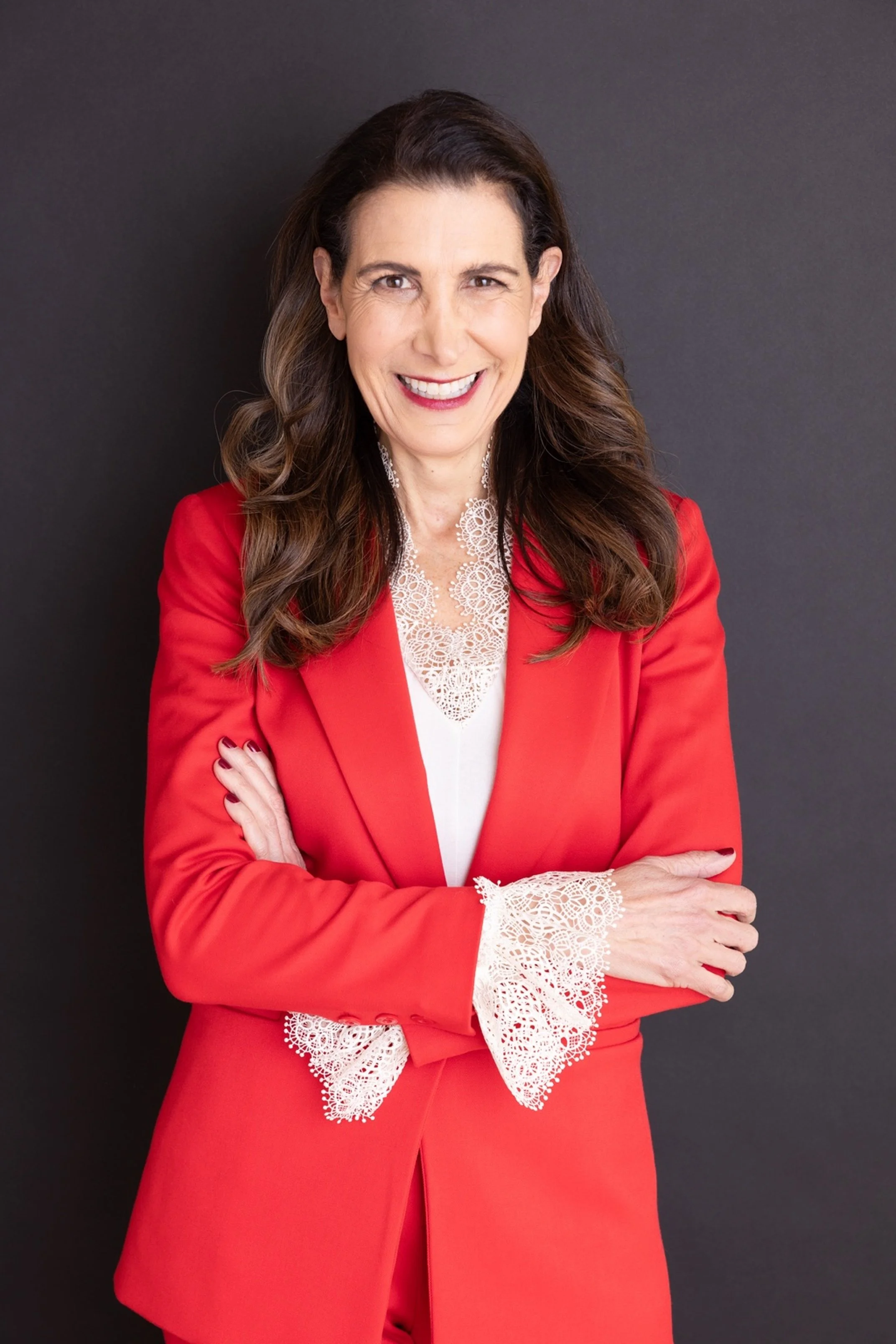 Tina Sharkey with long, wavy brown hair wearing a red blazer with lace cuffs over a white blouse, smiling and standing with arms crossed against a dark background.