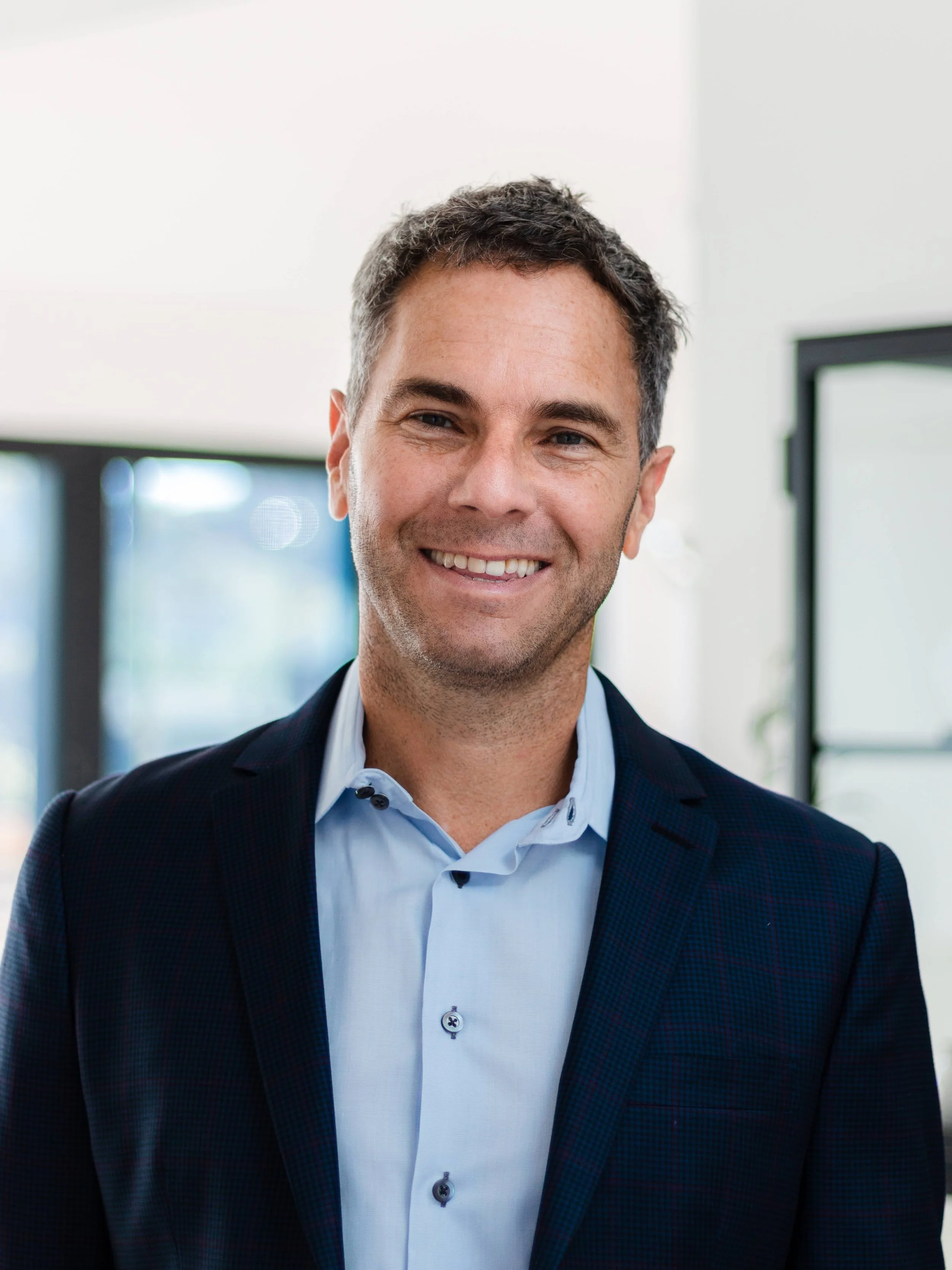 A smiling man in a suit with a blue shirt, standing indoors with natural light and windows in the background.