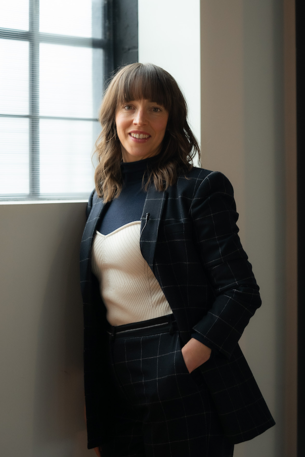A woman standing by a window with natural light, wearing a dark checked blazer and matching pants, with a white ribbed top, smiling at the camera.