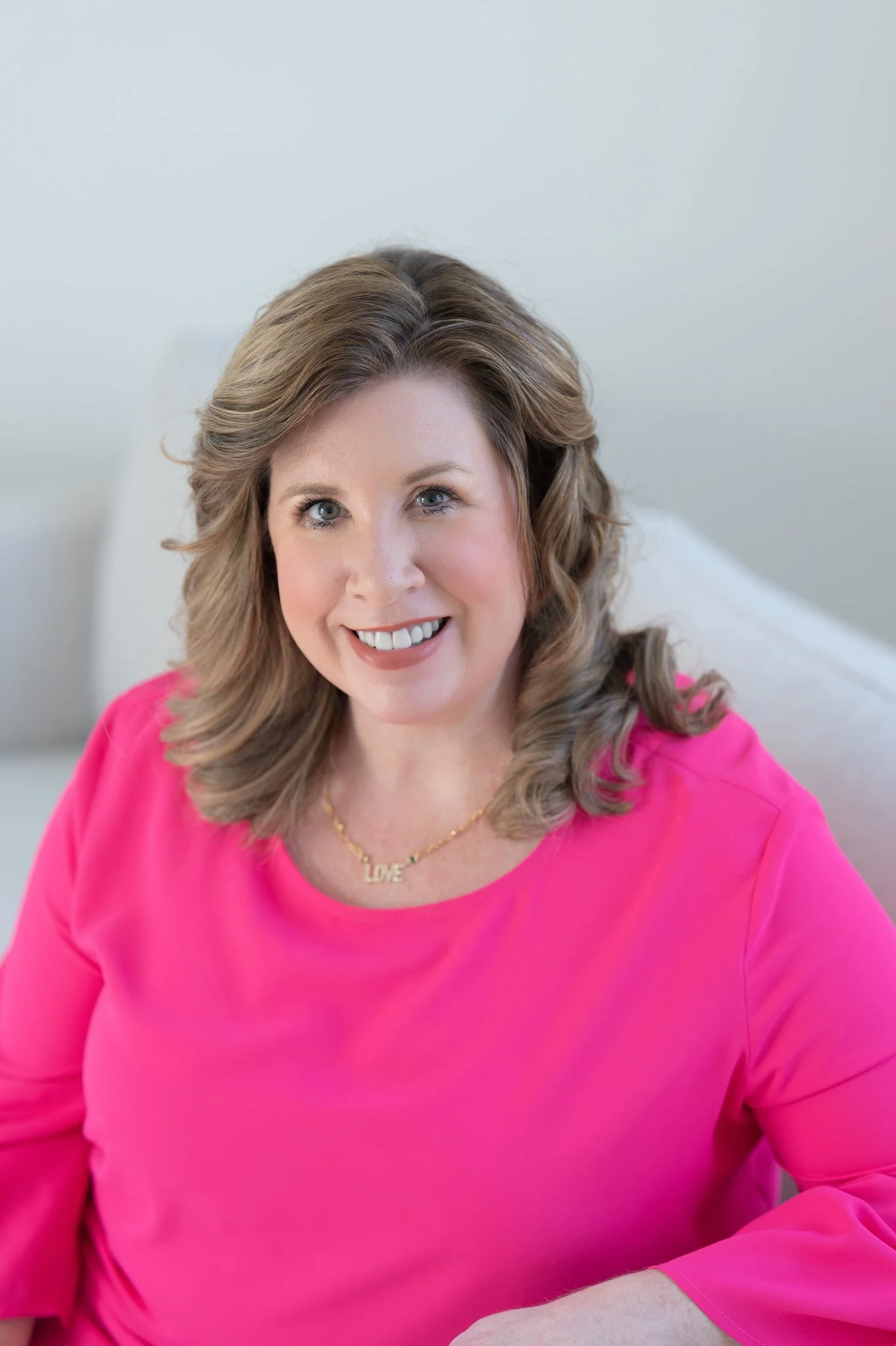 Caroline Maguire with shoulder-length, wavy brown hair, wearing a bright pink top and a gold necklace with 'LOVE' on it, smiling while sitting on a light-colored sofa in a neutral room.