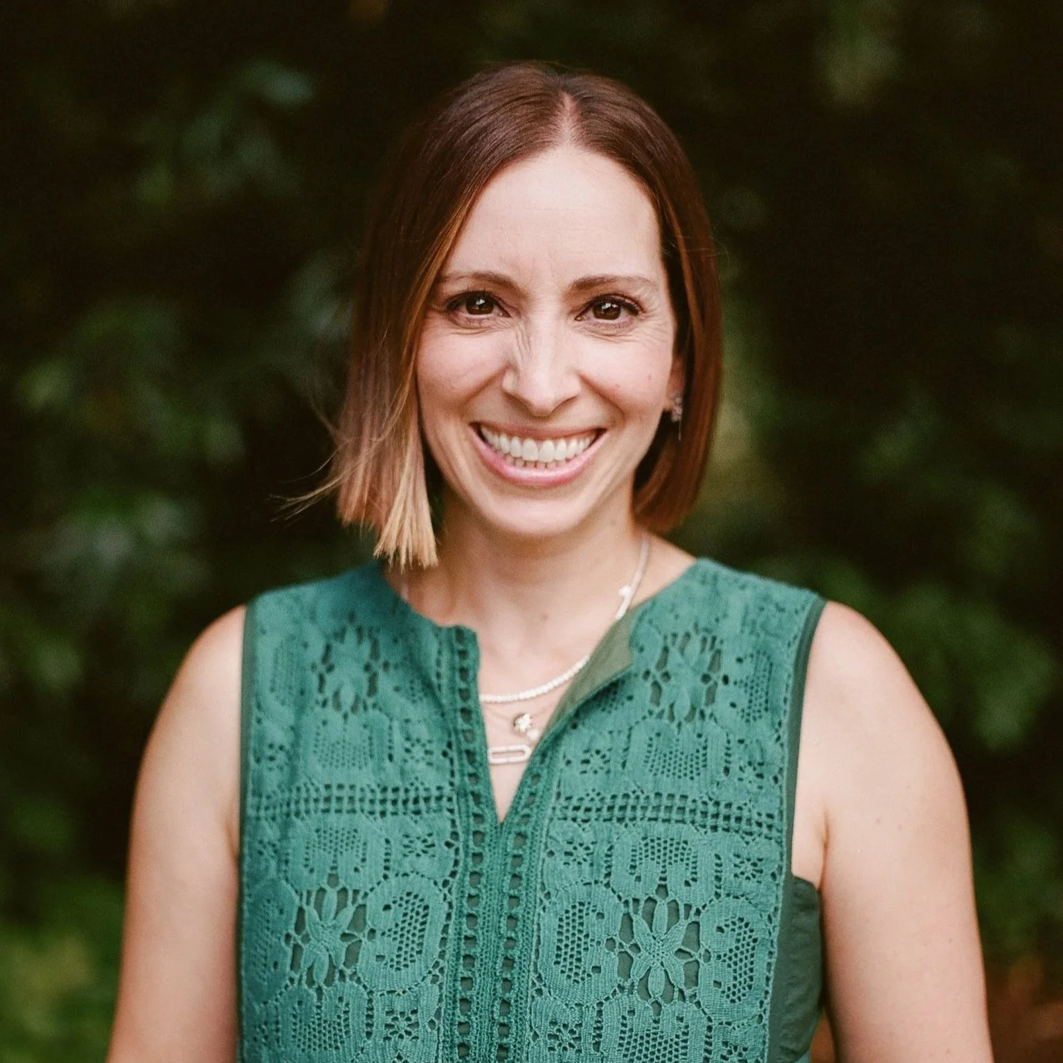 A woman with shoulder-length brown hair smiling outdoors, wearing a sleeveless green lace top and a pearl necklace, with a blurred green foliage background.