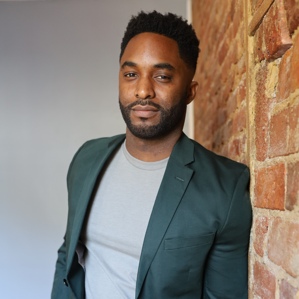 A man with dark curly hair, beard, and brown skin leaning against a brick wall, wearing a dark green blazer over a light gray T-shirt.