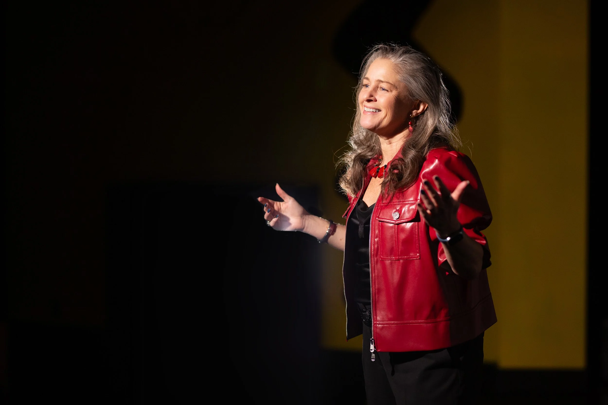 A woman with gray hair wearing a red leather jacket and black clothing, standing on stage with a smile and open arms, speaking or presenting.