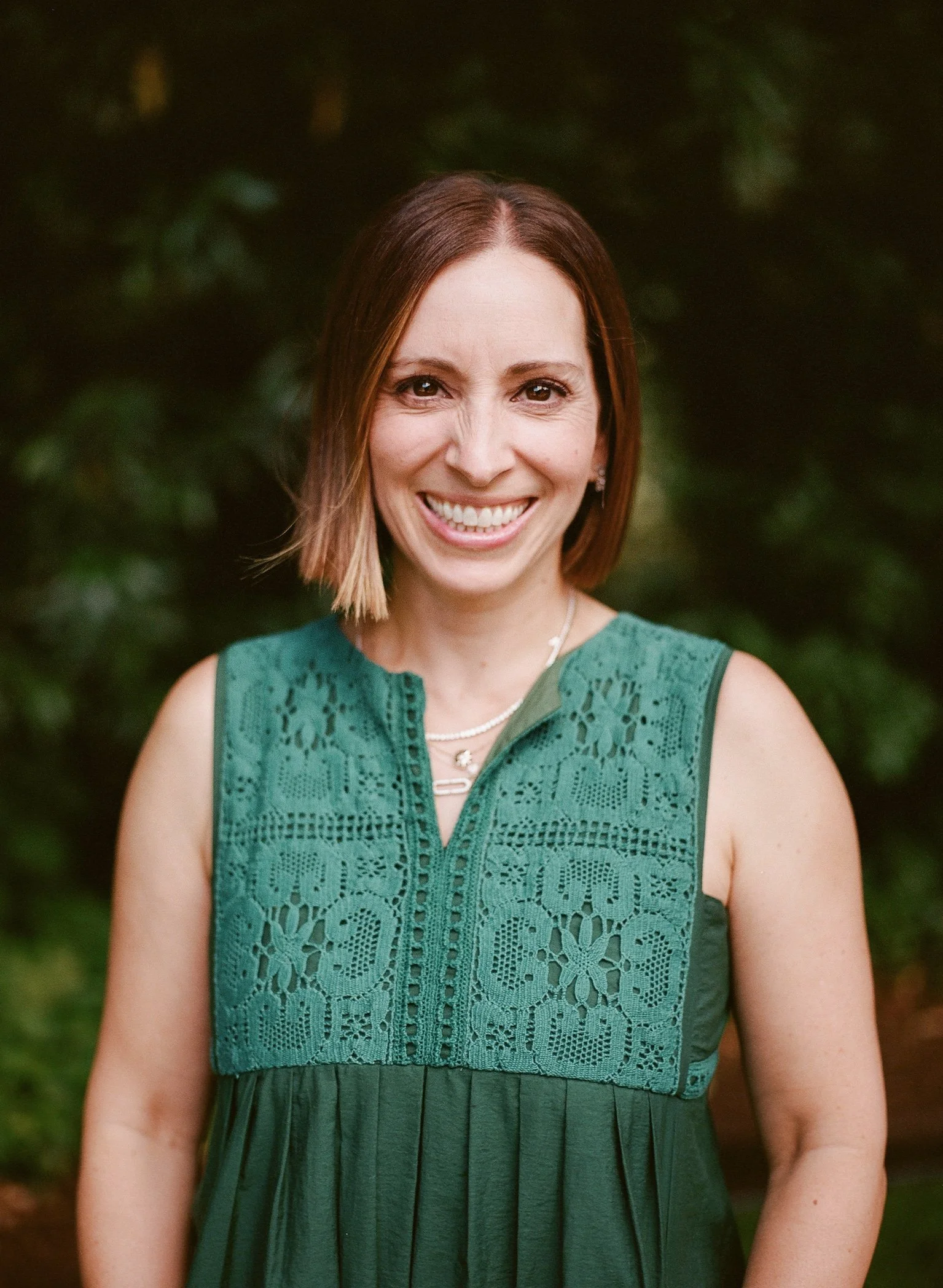 A smiling woman with shoulder-length brown hair, wearing a green sleeveless dress with lace detailing on the top, standing outdoors with a blurred, dark green background.