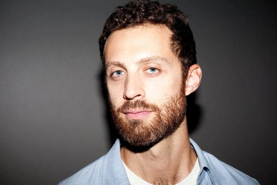 Close-up of a man with curly brown hair, blue eyes, and a beard, wearing a light blue shirt against a dark background.