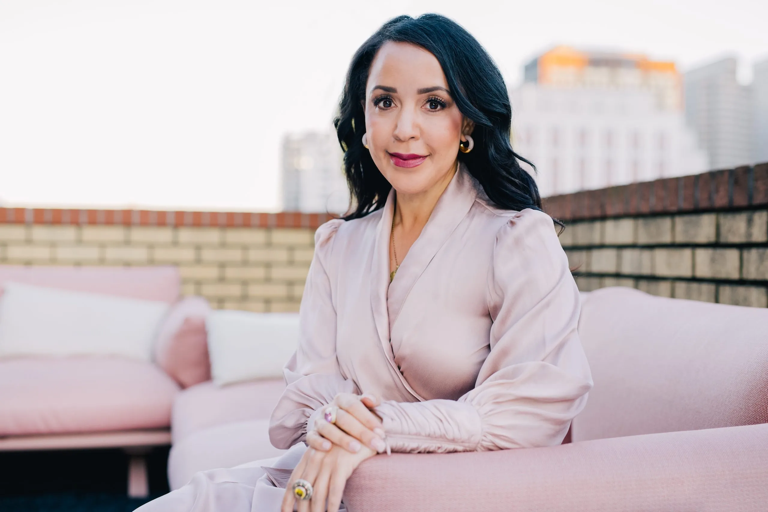 Julia Collins with black hair and gold jewelry sitting on a pink outdoor couch on a rooftop with city buildings in the background.