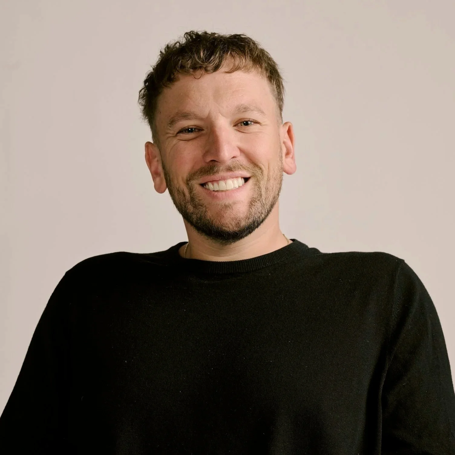 A young man with short curly hair, a beard, and a big smile, wearing a black shirt, standing against a plain background.