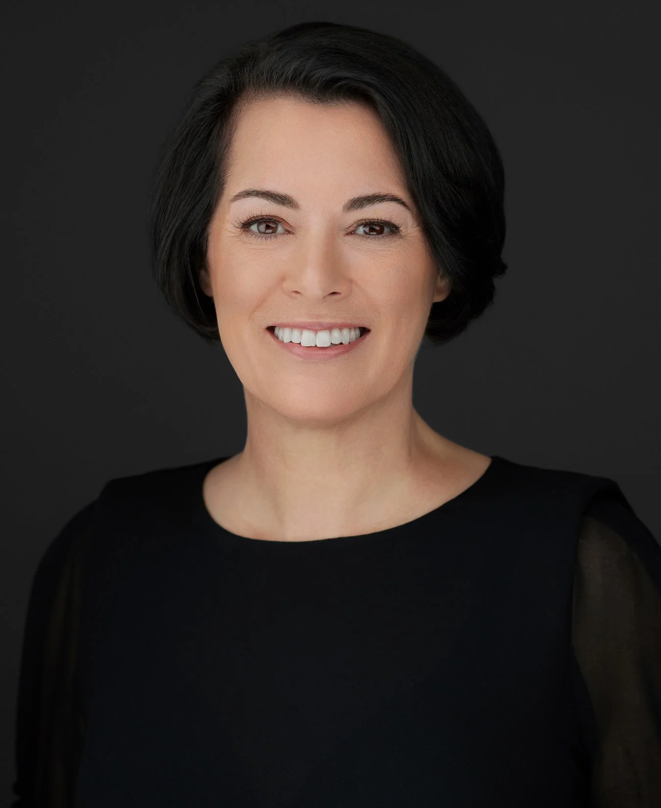 Nicole Malakowski with short black hair, smiling, wearing a black top, against a dark background.