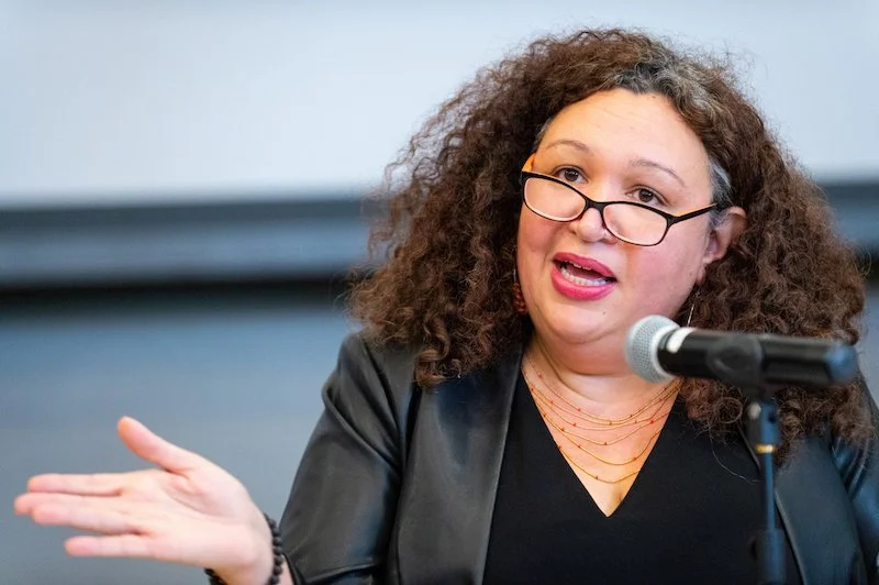 A woman with curly brown hair and glasses speaking into a microphone, gesturing with her hand.