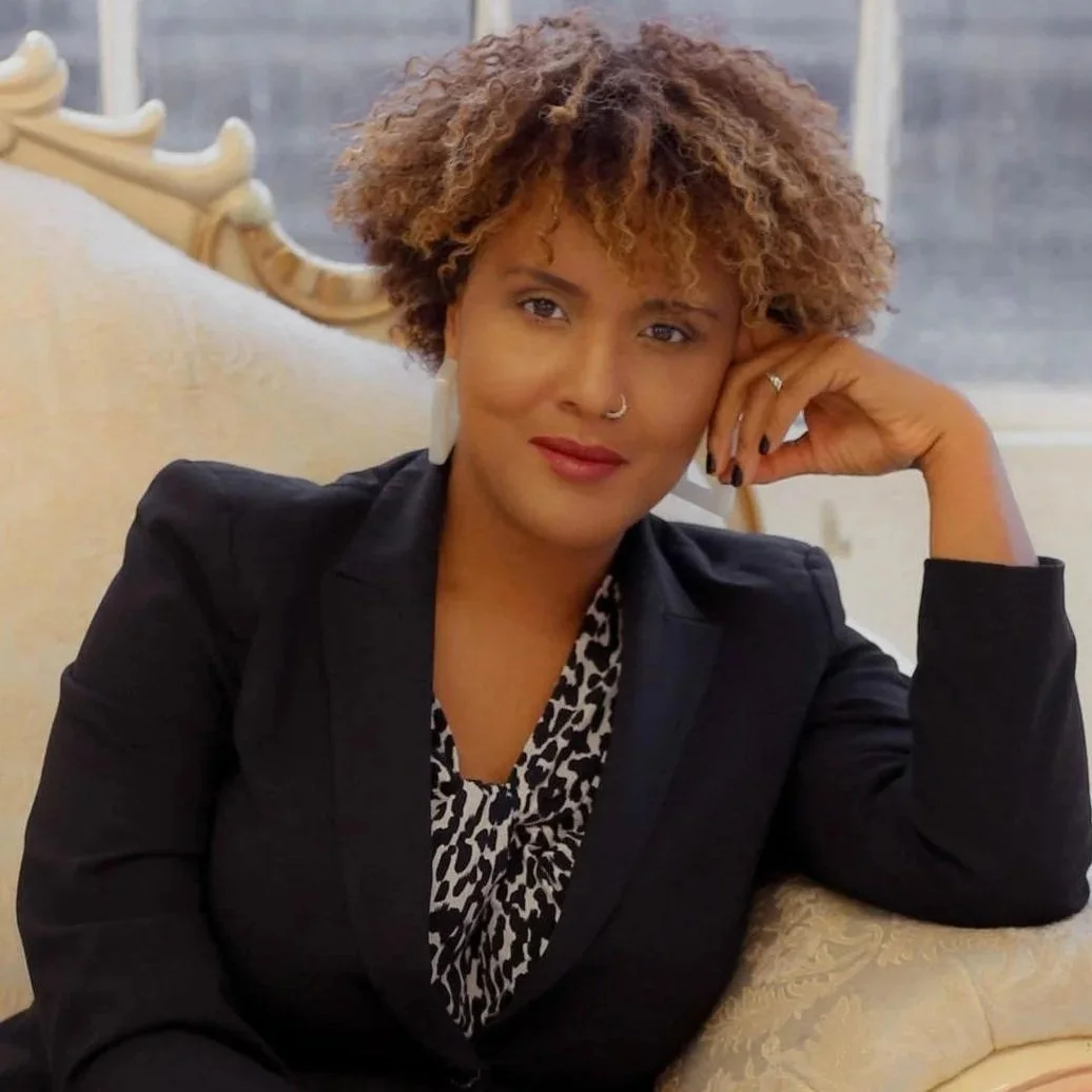 Crystal Fleming, with short, curly hair and light brown skin, sitting on a beige sofa, wearing a black blazer and a patterned top, with her hand resting on her head.