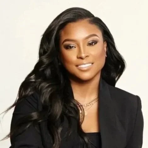 Portrait of a smiling woman with long, wavy black hair, wearing a black blazer and gold jewelry against a light background.