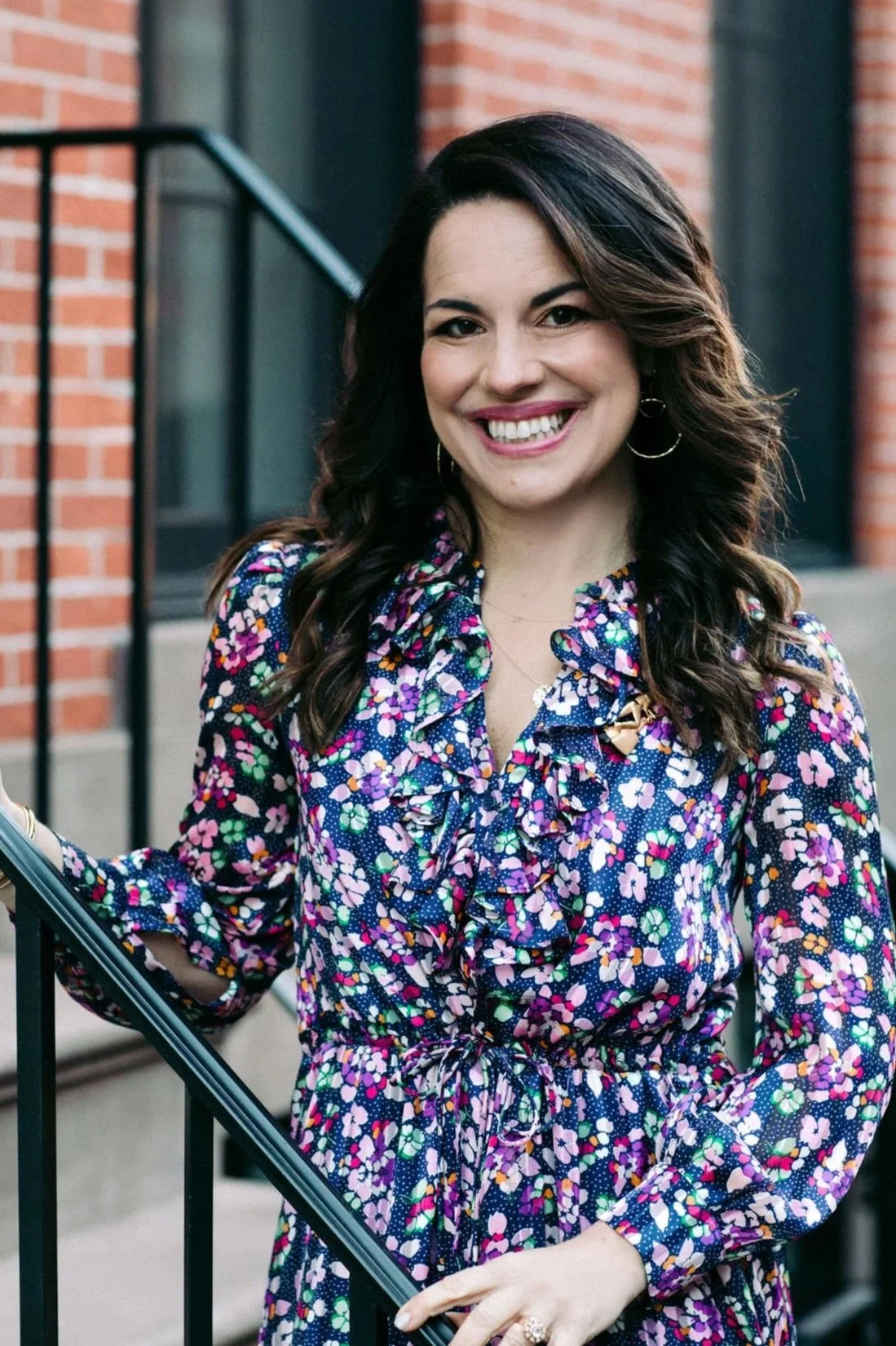 A woman with dark, wavy hair smiling, wearing a colorful floral dress with ruffles, standing outside near a staircase with a brick building in the background.