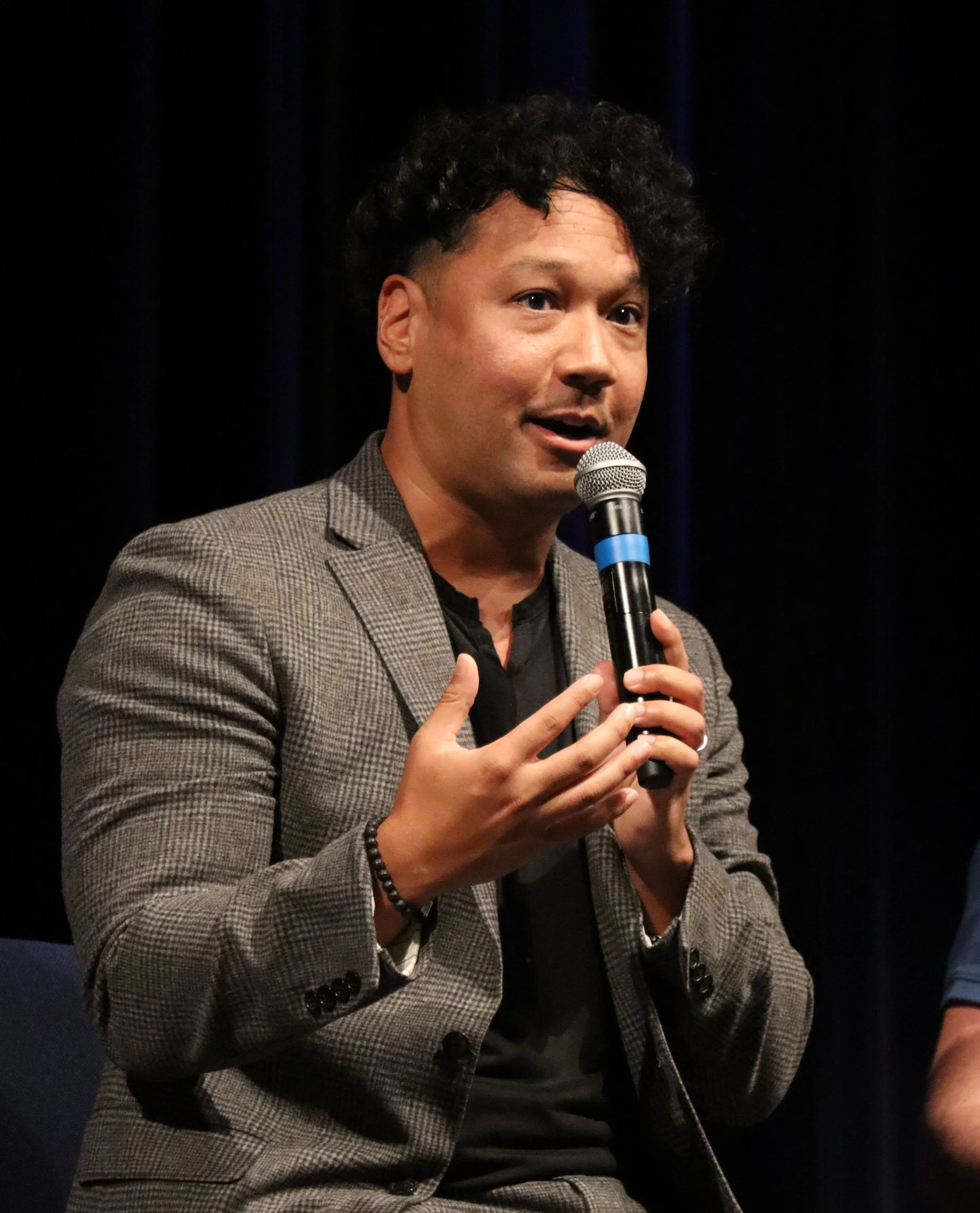Brad Jenkins speaking into a microphone during a presentation, wearing a checkered blazer and black shirt, with dark curly hair, seated against a dark background.
