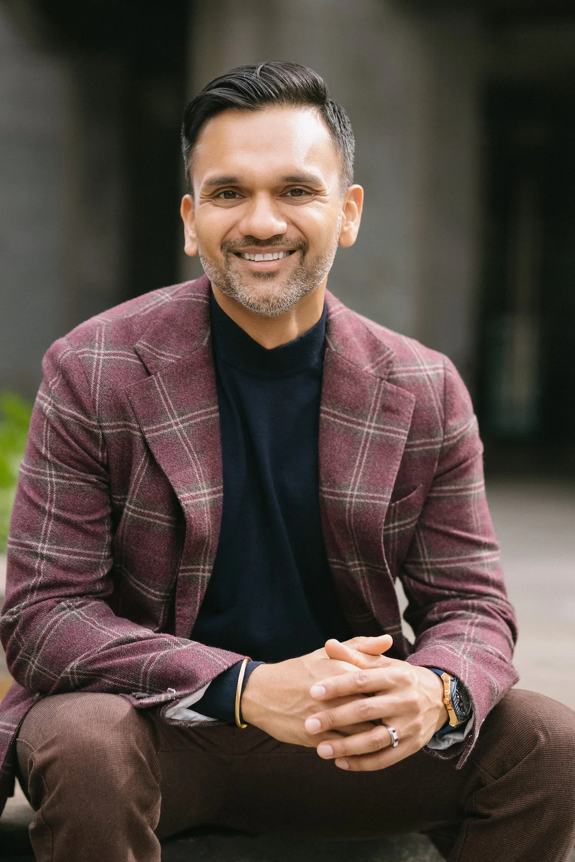 A smiling Anu Gupta sitting outdoors, wearing a burgundy plaid blazer, black turtleneck, and brown pants.