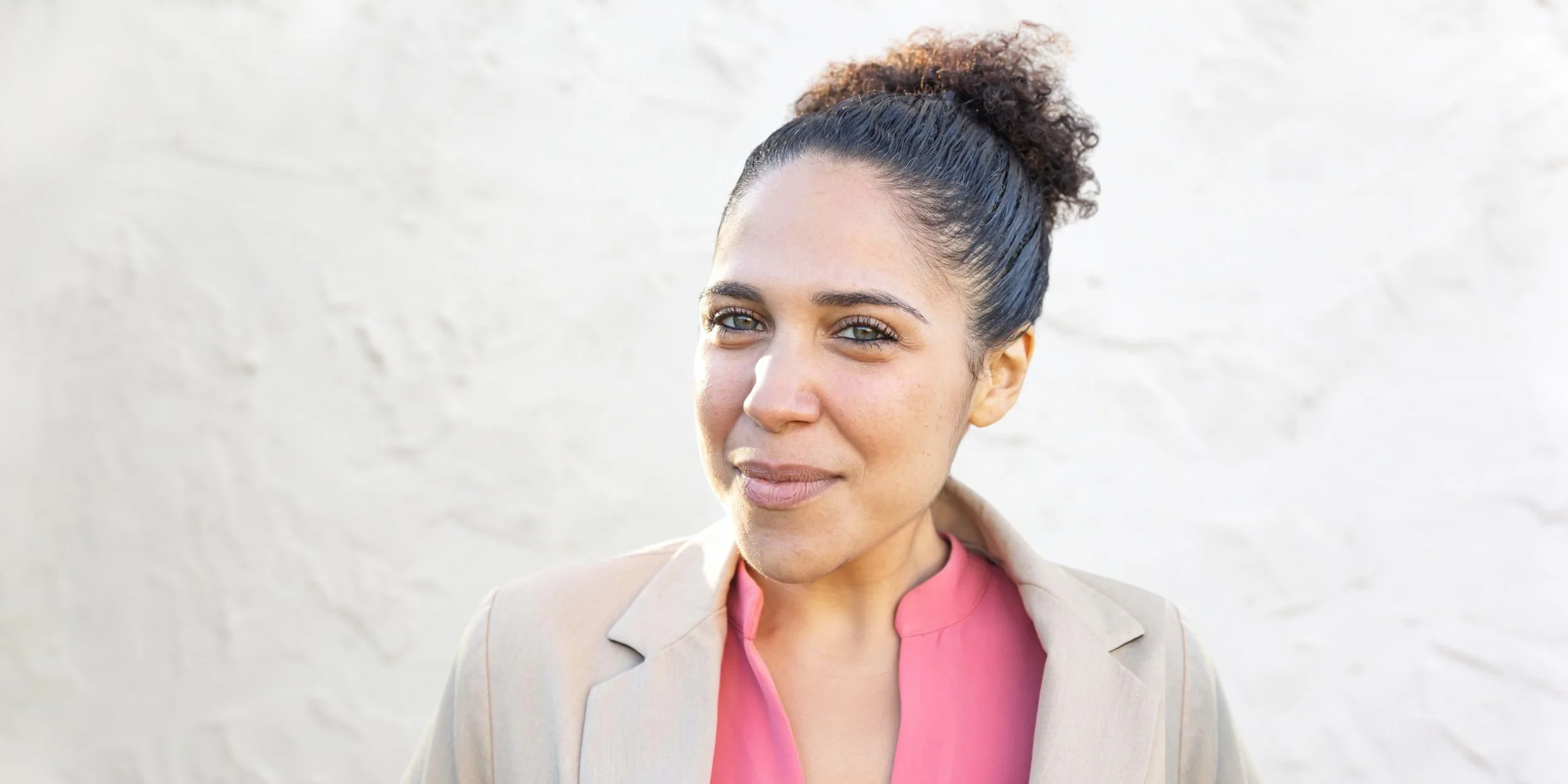 A woman with curly hair tied back, wearing a beige blazer and pink shirt, smiling against a light-colored textured wall.