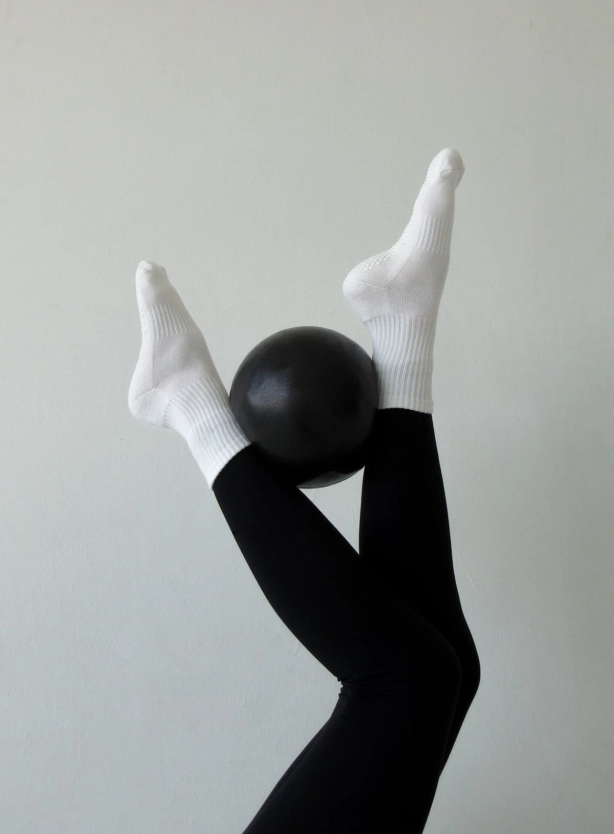 Person wearing black leggings and white socks balancing a black fitness ball on their feet against a plain light background.