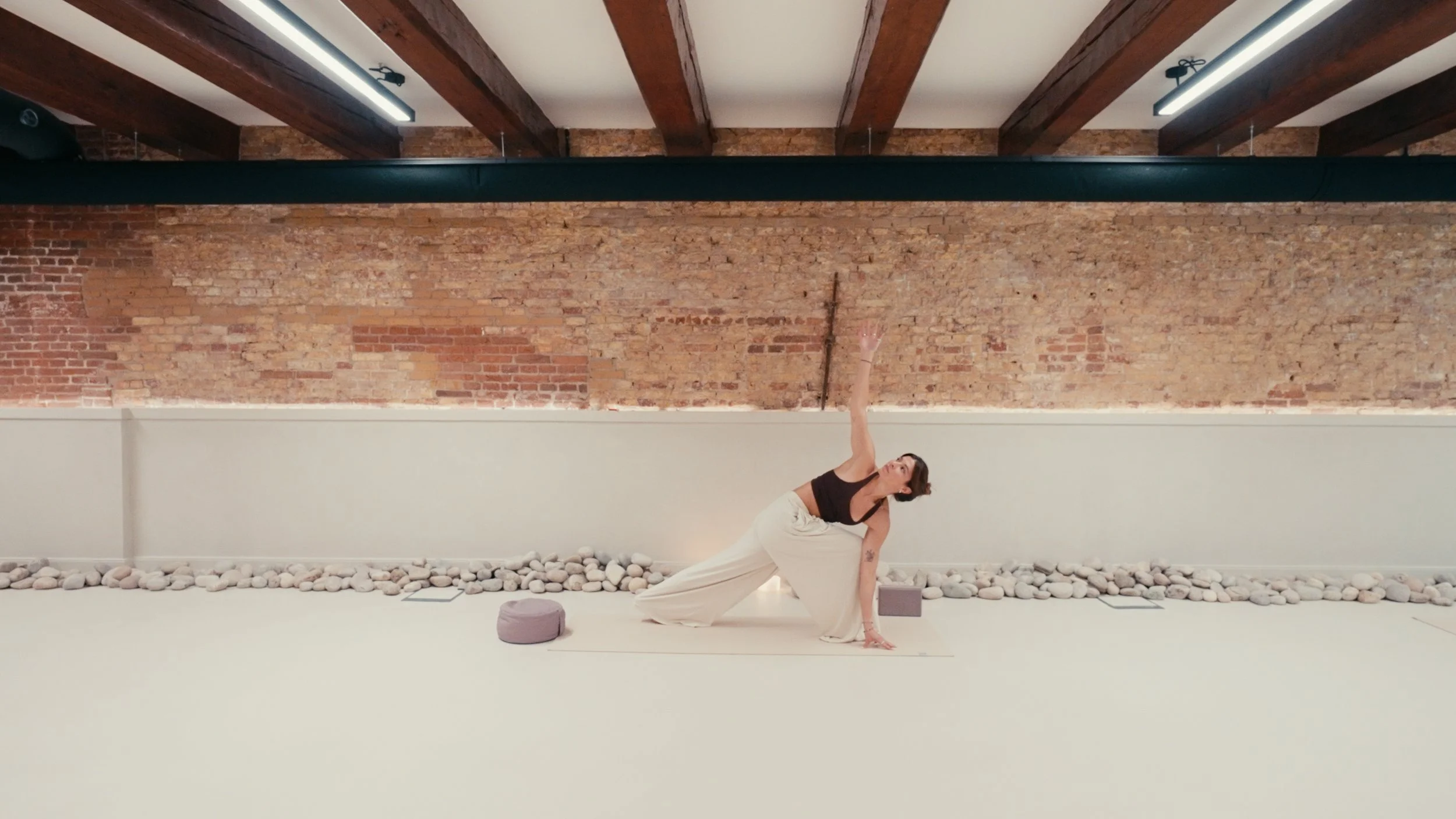 A woman practicing yoga in a minimalist studio with a brick wall and wooden beams. She is in a side stretch pose, wearing a black top and beige pants.