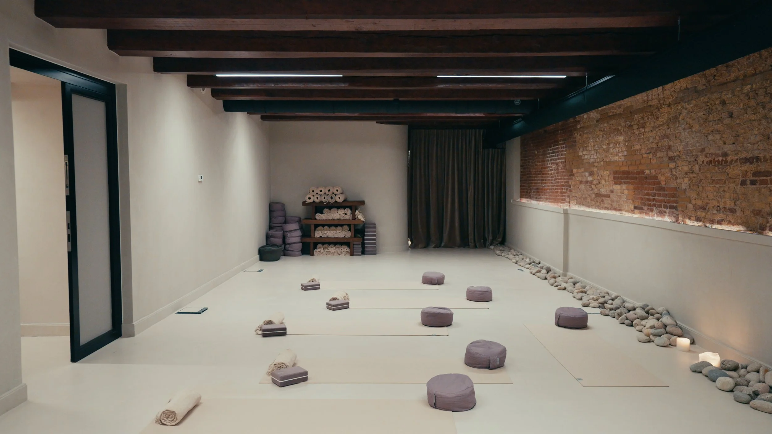 Empty yoga or meditation studio with beige mats, cushioned blocks, and round cushions on the floor, stacked towels and yoga props on shelves, exposed brick wall, and ambient lighting.