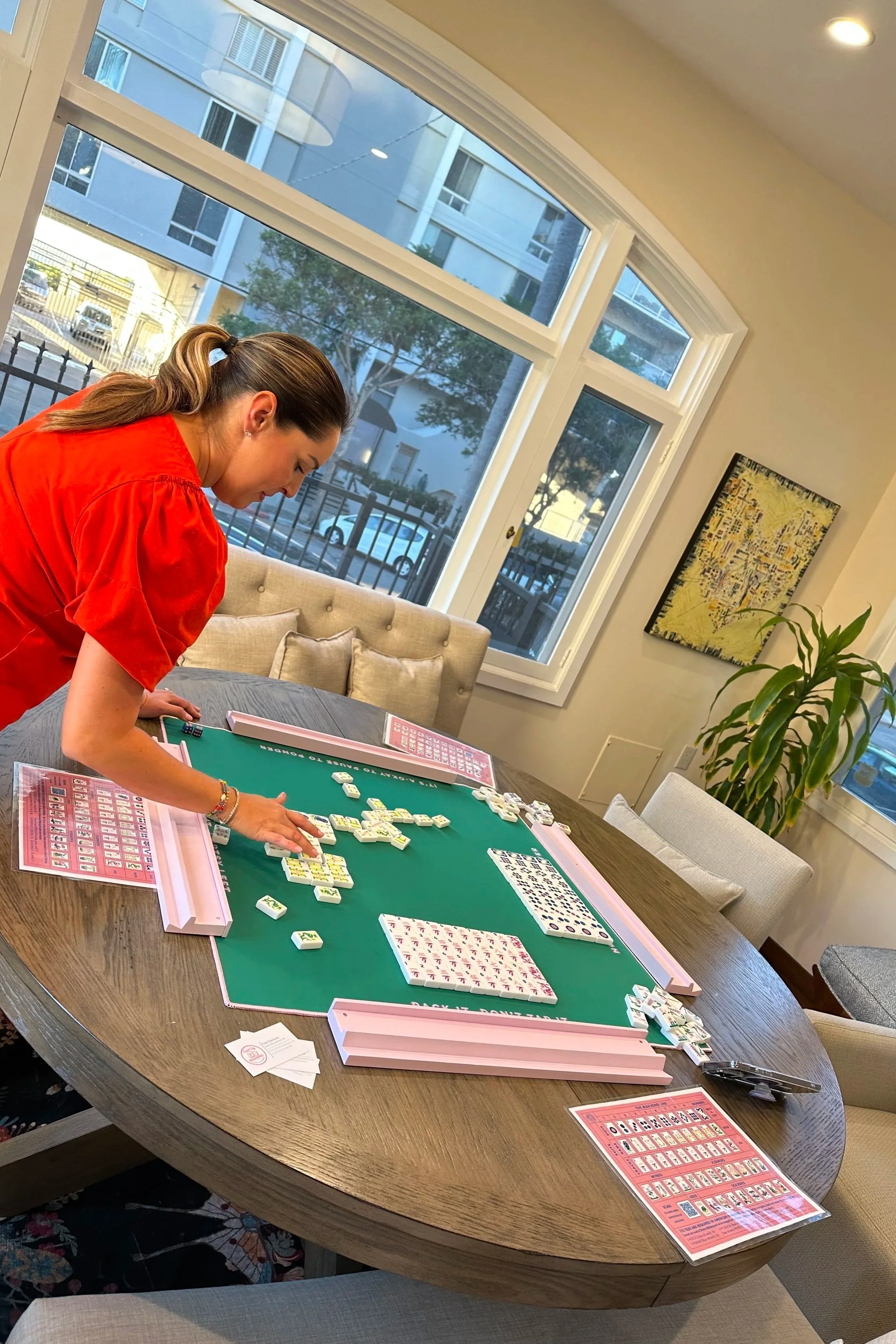 A woman in a red shirt playing Mahjong at a wooden table in a bright room with large windows, beige chairs, and a potted plant.