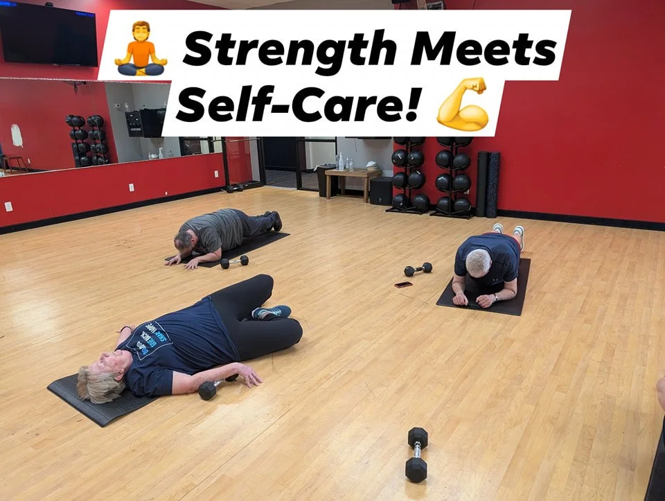 People doing floor exercises with dumbbells in a fitness studio with a motivational sign that says 'Strength Meets Self-Care!'