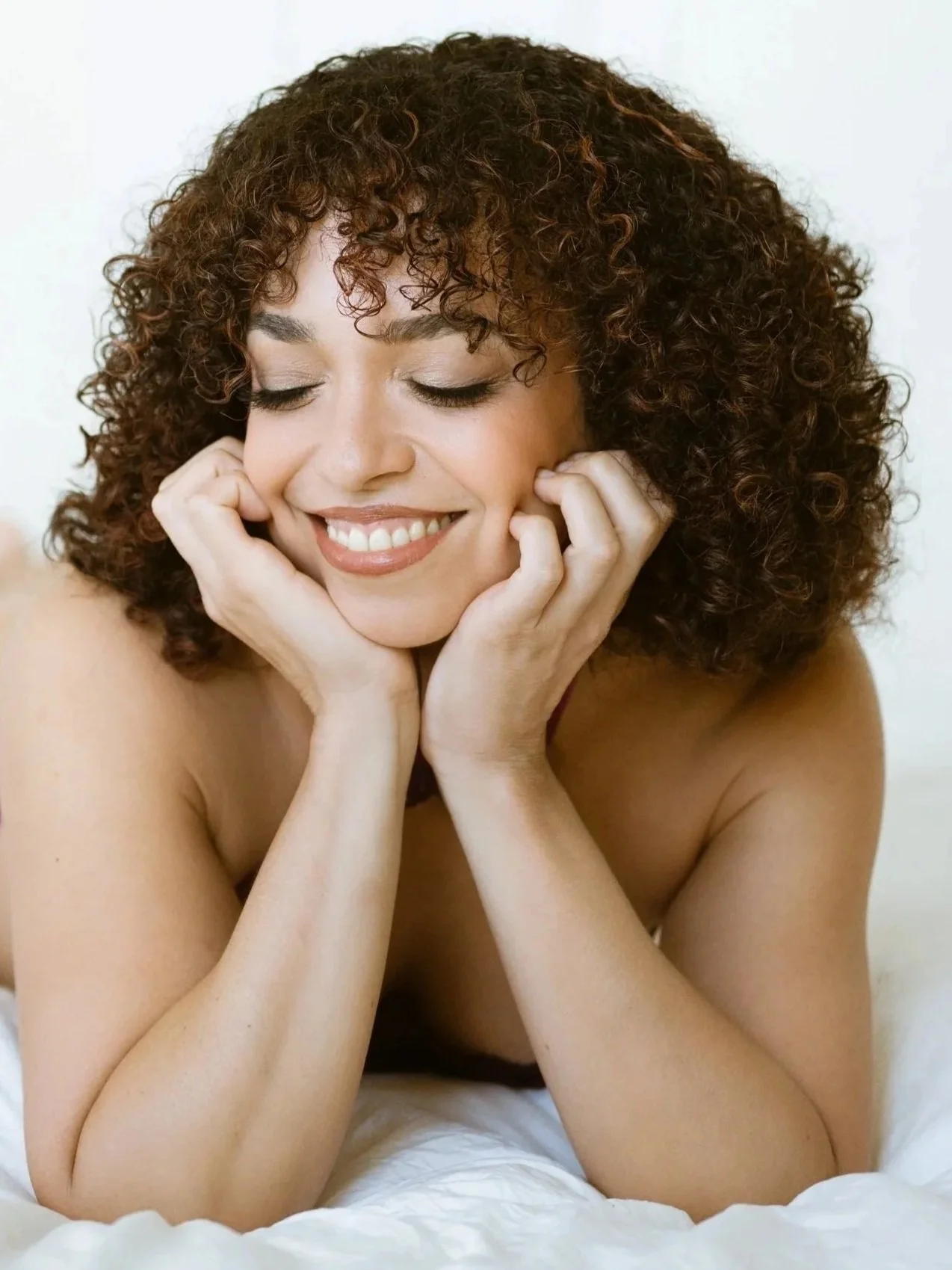 A woman with curly brown hair smiling with her eyes closed, resting her face in her hands, in a relaxed pose.