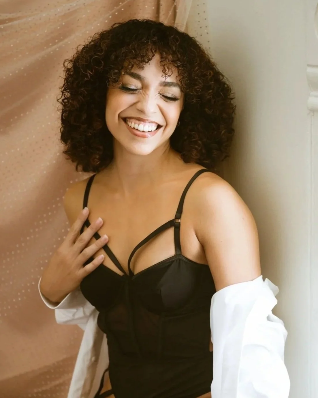 A woman with curly hair, smiling with eyes closed, wearing black top and a white shirt falling off her shoulders, standing by a beige curtain and light-colored wall.