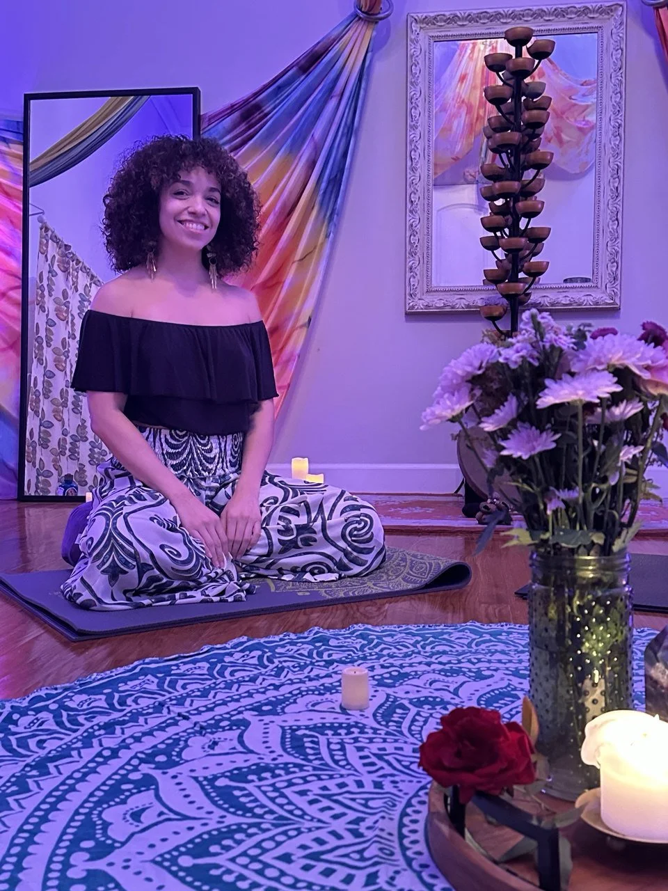 A woman with curly hair smiling, sitting on a yoga mat in a room decorated for a ceremony with candles, flowers, a mirror, and colorful drapery.