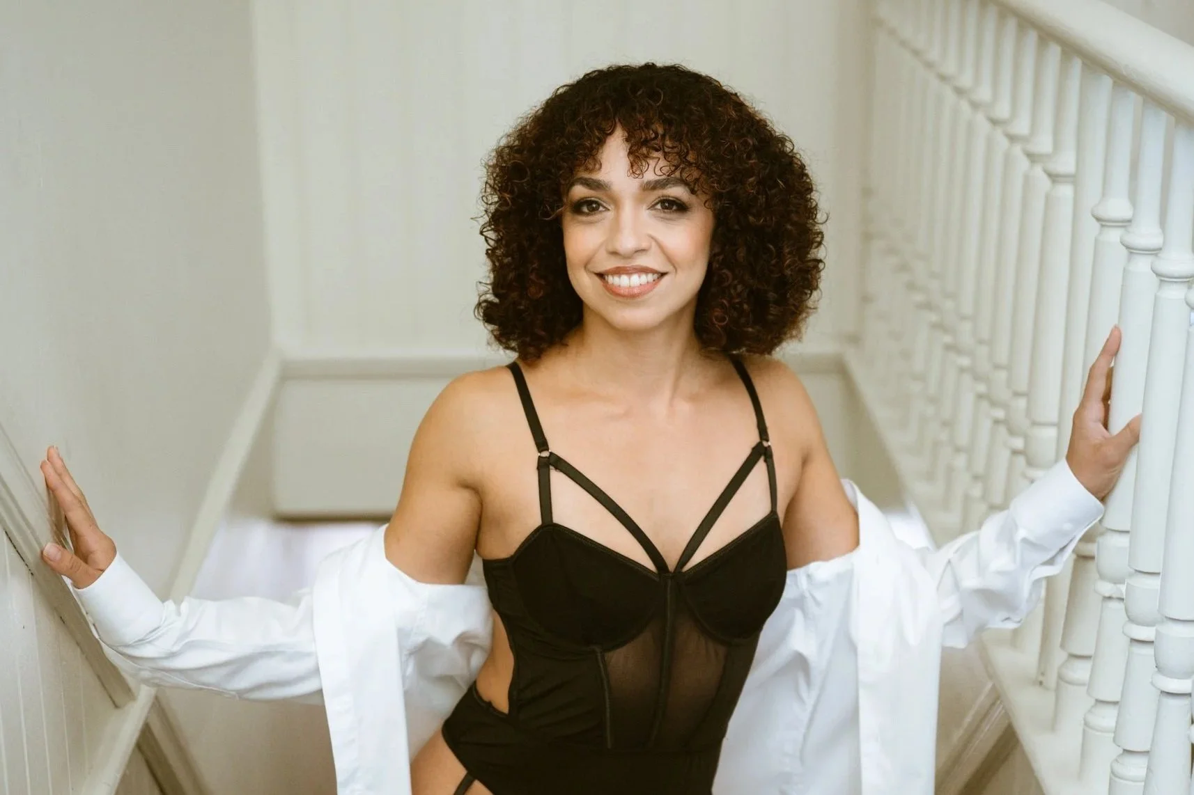 A woman with curly brown hair smiling, wearing a black top and a white shirt draped over her shoulders, going up a staircase.