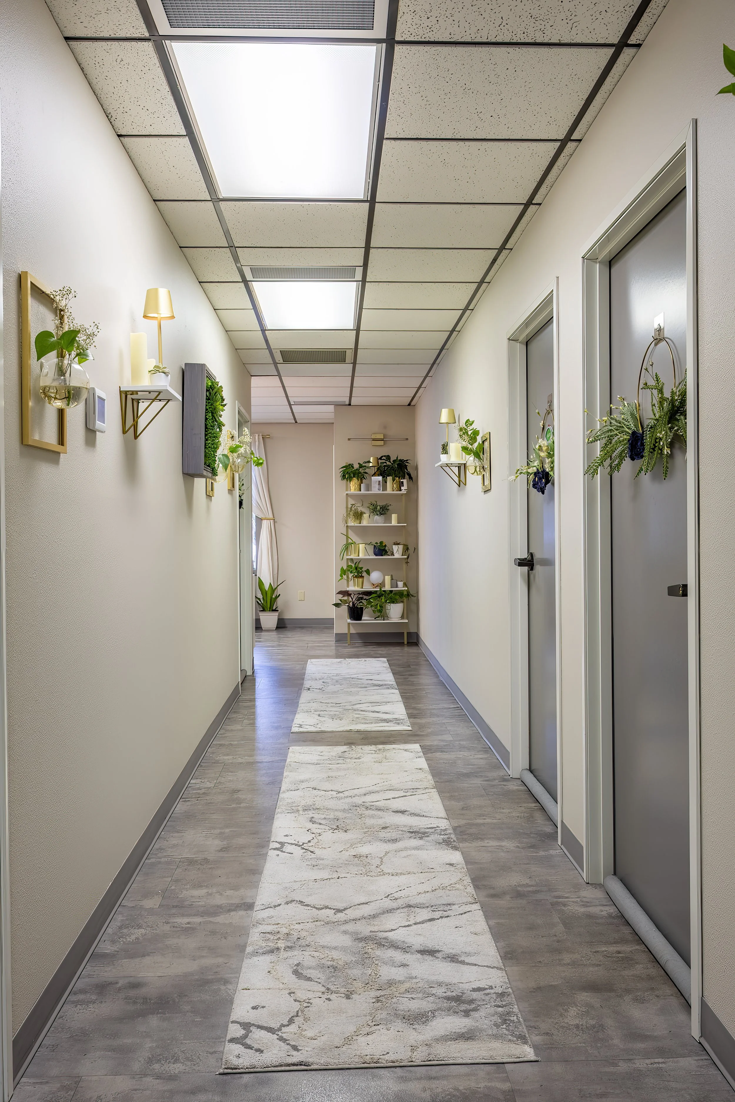 A hallway with beige walls, gray doors decorated with flower and greenery wreaths, a white marble runner rug, and wall-mounted lights with plant accents. There is a white shelf with potted plants at the end of the hall.