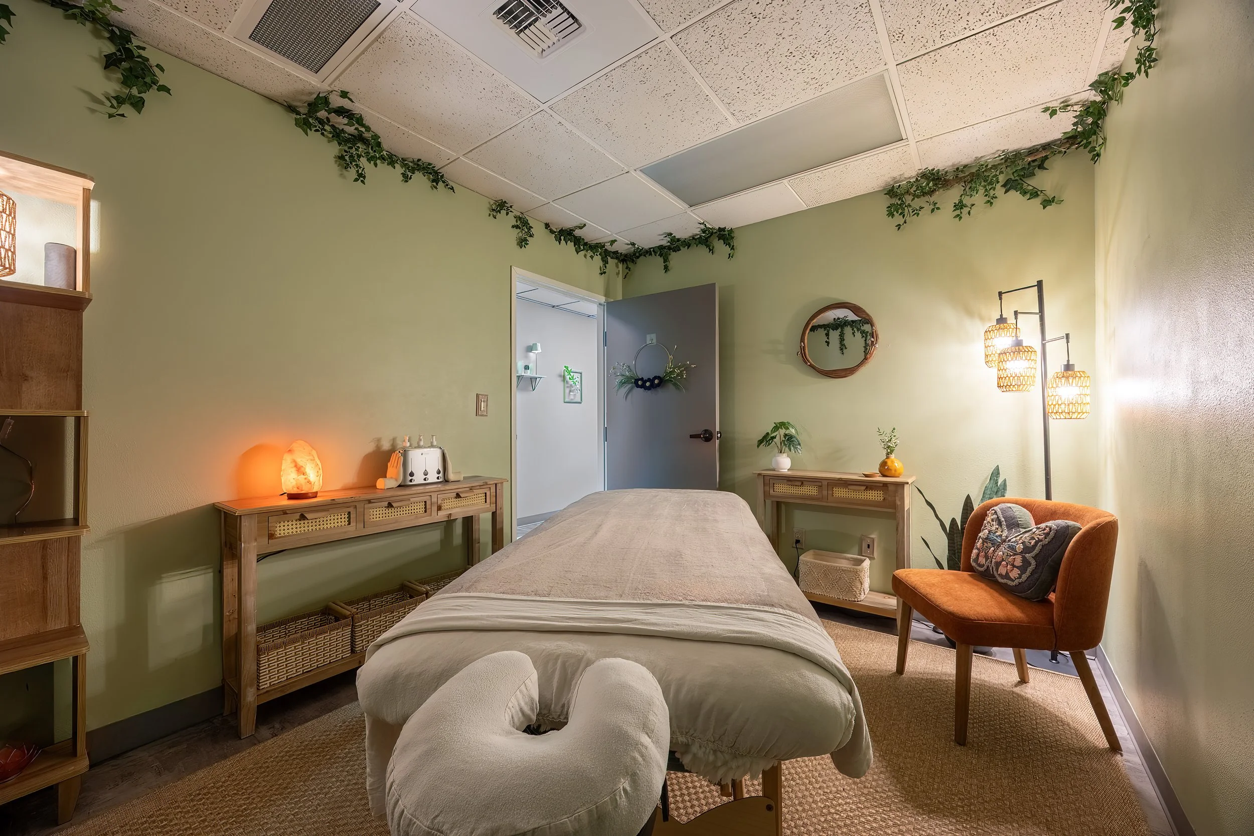A calming massage therapy room with a massage table in the center, warm lighting, green walls decorated with plants and leaf designs, and cozy furniture.