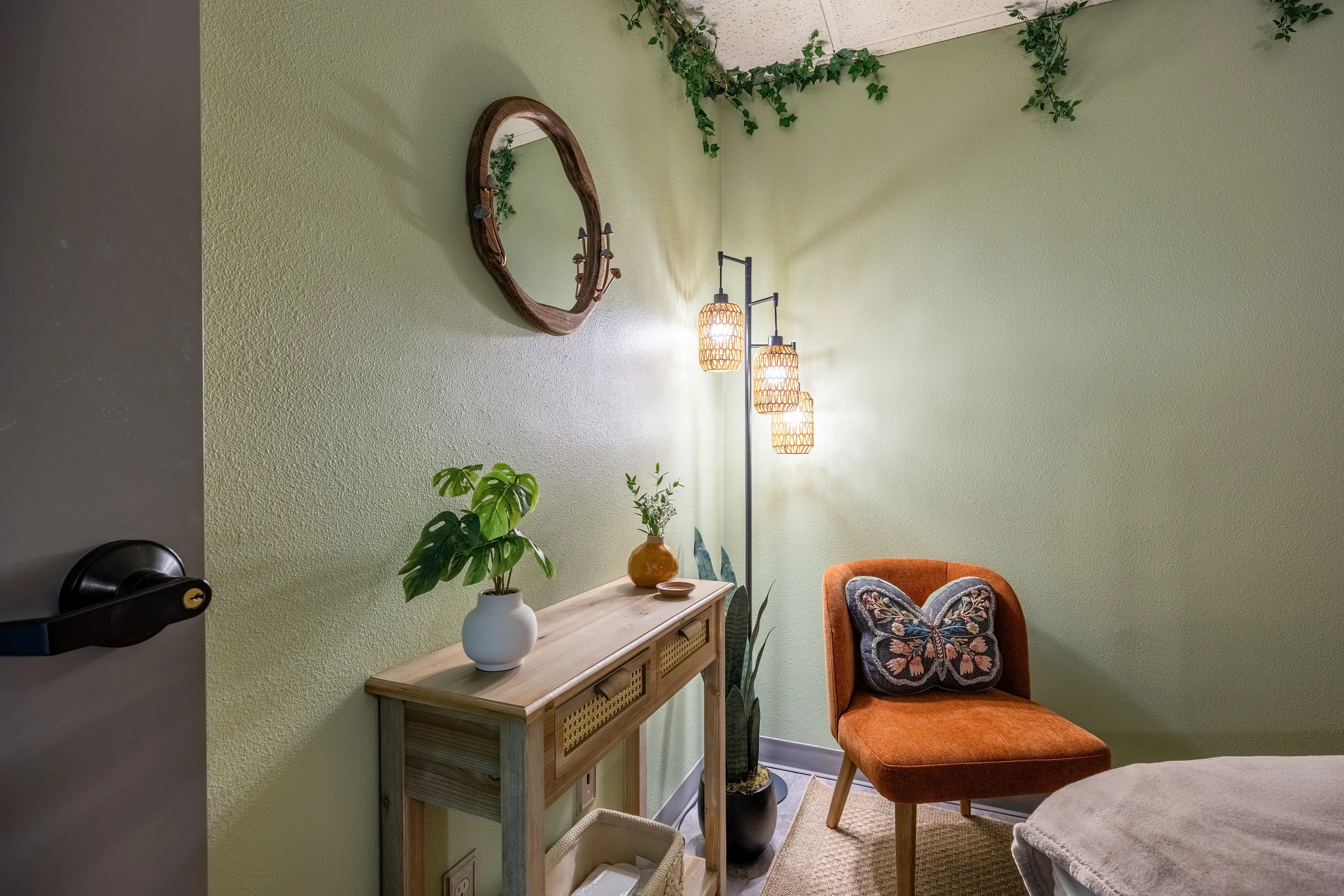 Corner of a room with light green walls, a wooden console table with potted plants, a standing floor lamp with three woven pendant lights, a round mirror, and an orange chair with a butterfly-shaped pillow.