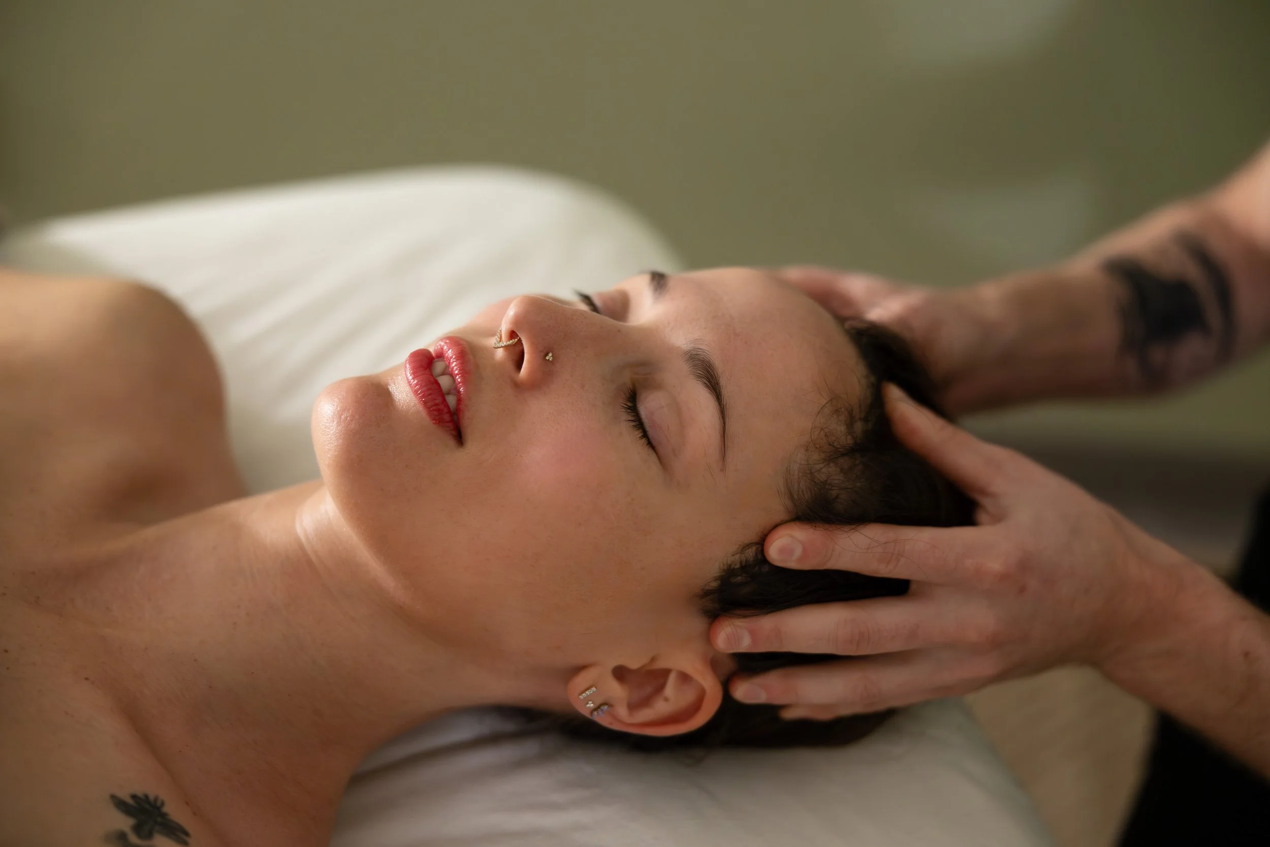 A woman with dark hair receiving a massage, lying on her back with eyes closed, in a calm environment.