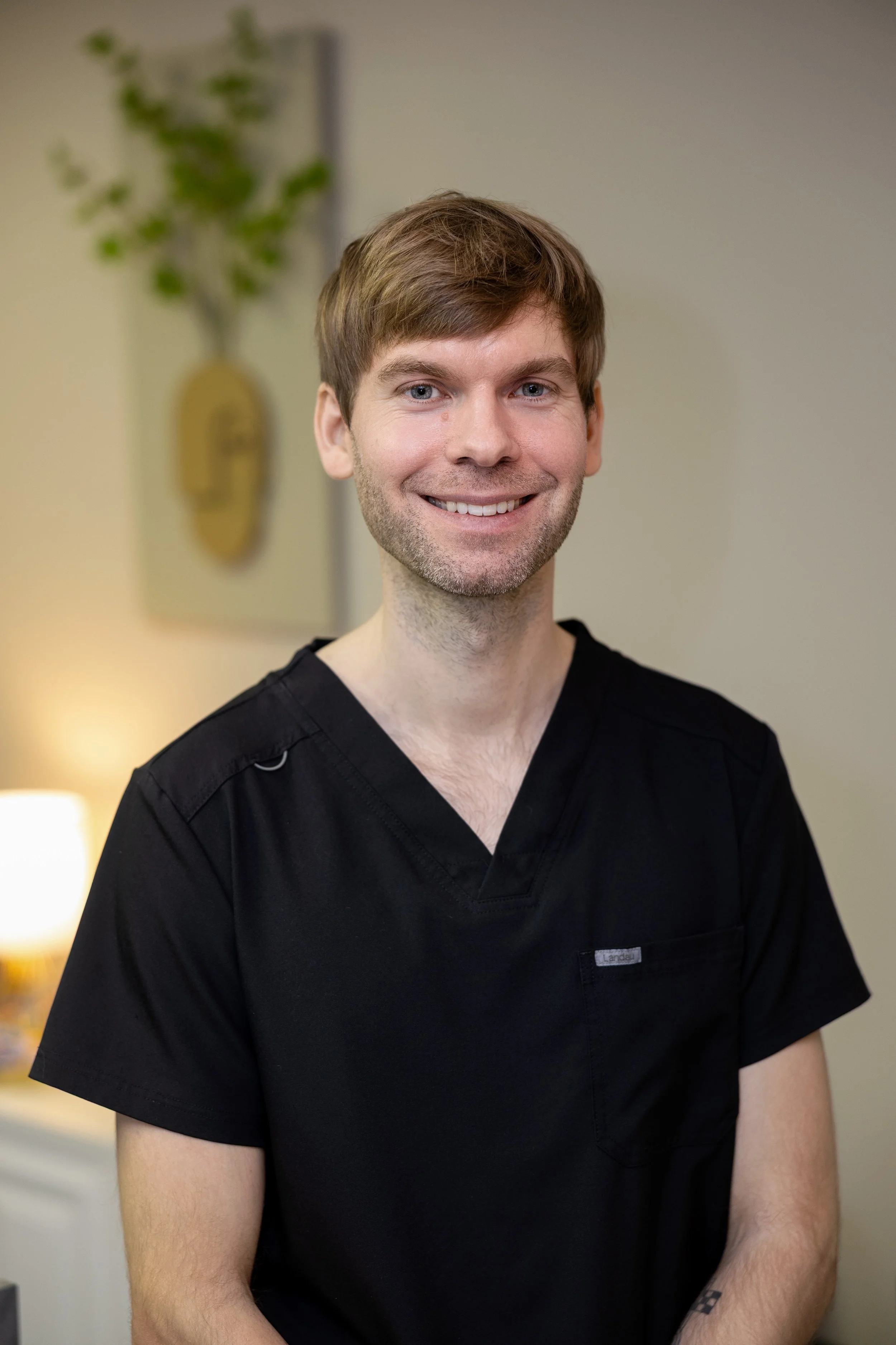 A smiling man with light brown hair in black scrubs standing indoors with a blurred background of a lamp and artwork on the wall.