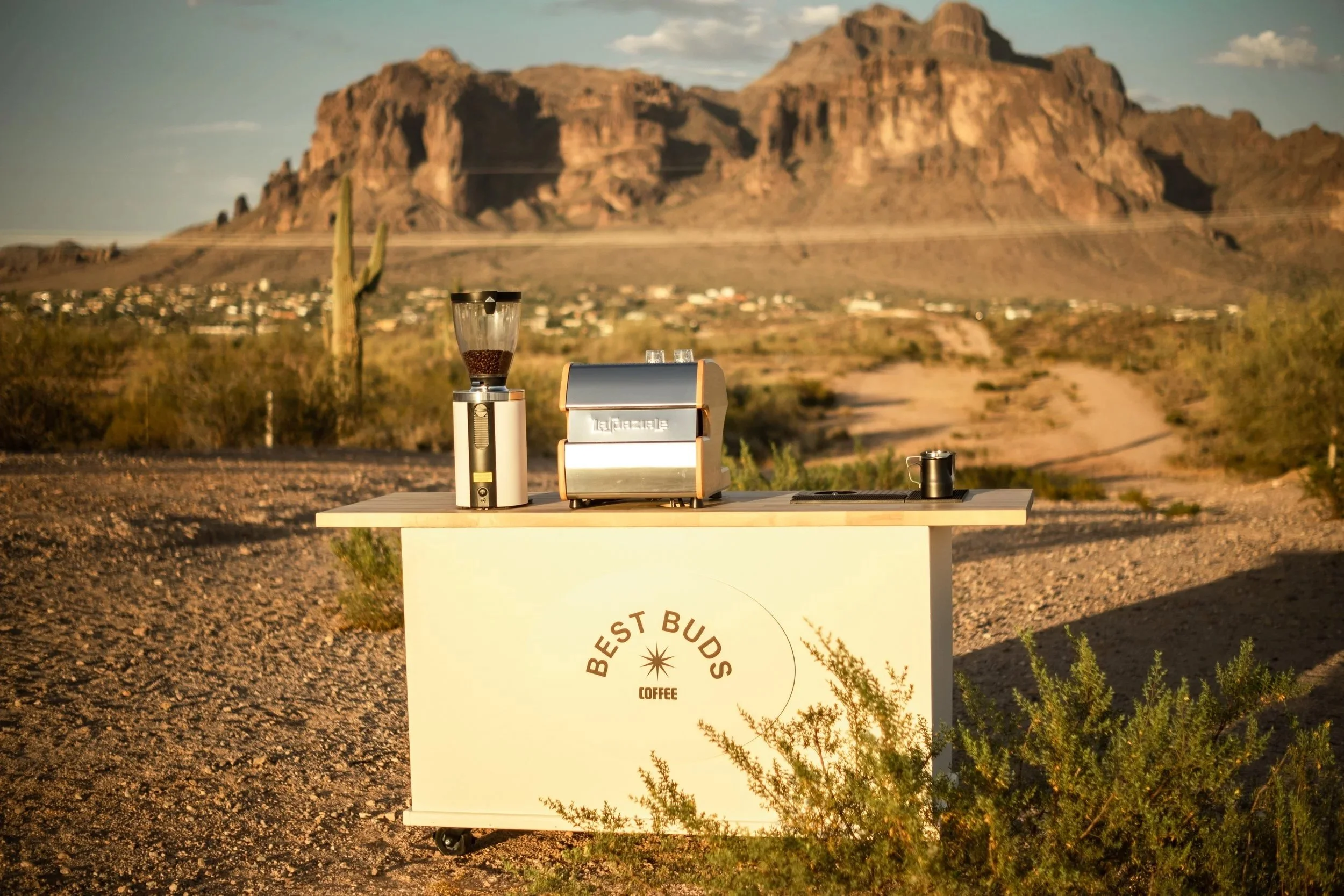 A table with a coffee grinder, a toast cutter, and a small pitcher set against a desert landscape with mountains and a cactus in the background.