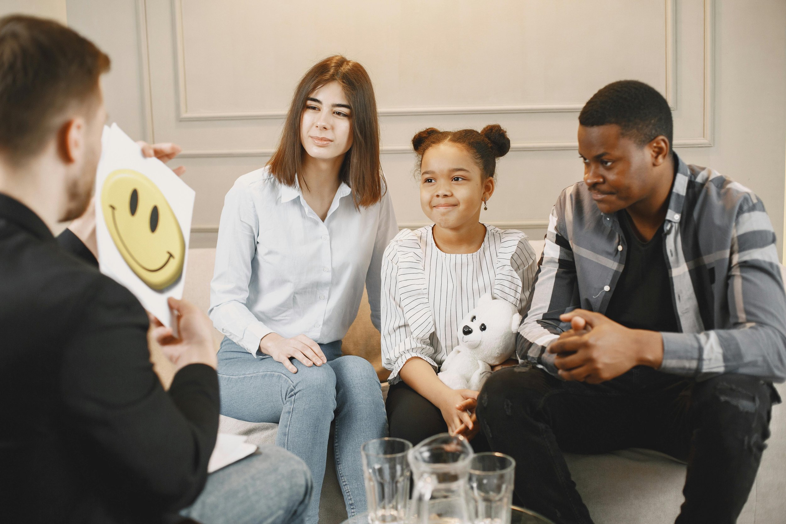 A group therapy session with four people, including a woman and a young girl holding a stuffed animal, listening to a person holding a paper with a smiley face.