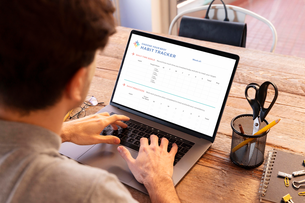 Person using a laptop to fill out a habit tracker on a wooden desk, with office supplies nearby.