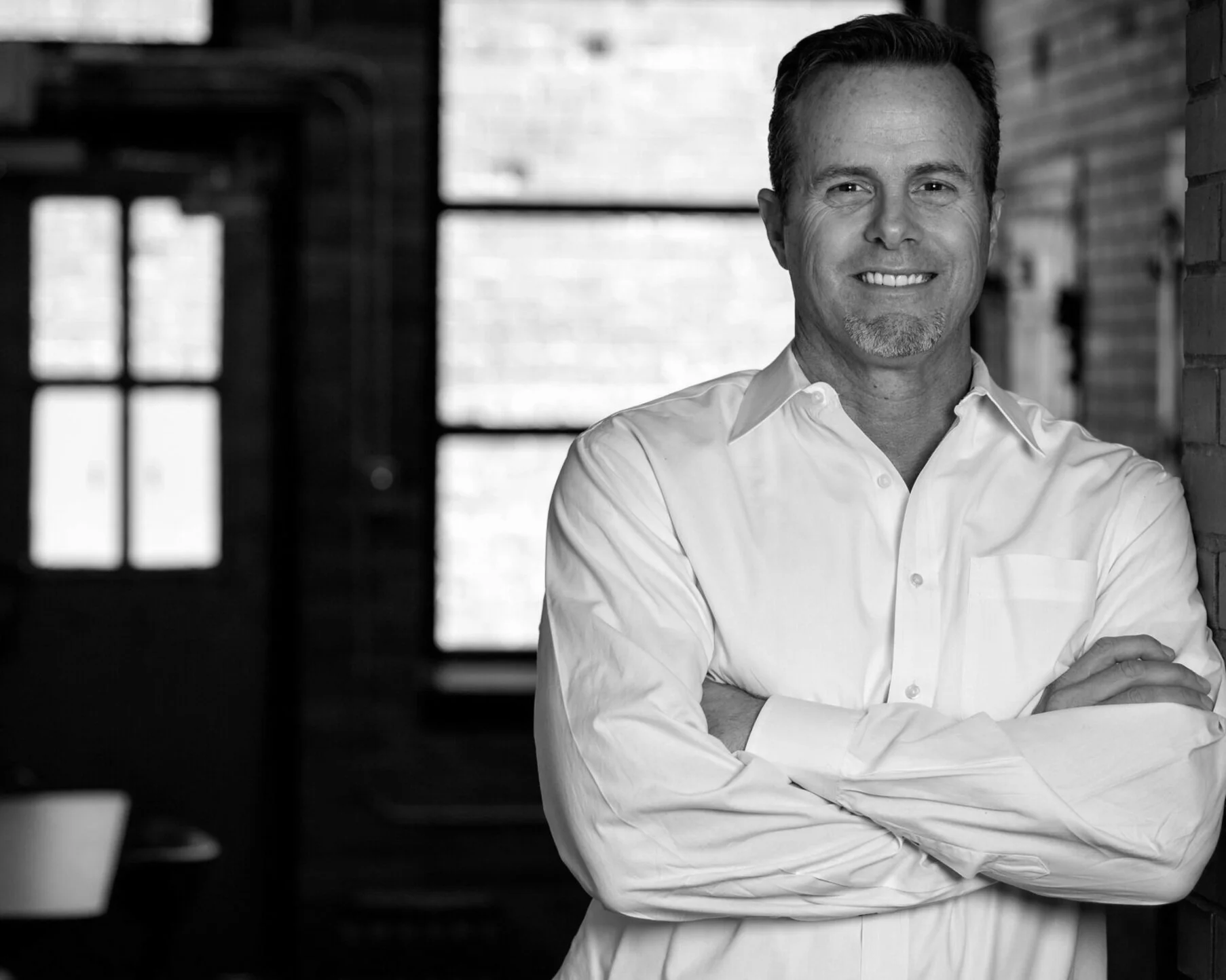 Black and white photo of a smiling man with short hair, wearing a white button-up shirt, standing with arms crossed in front of a brick wall and large windows.
