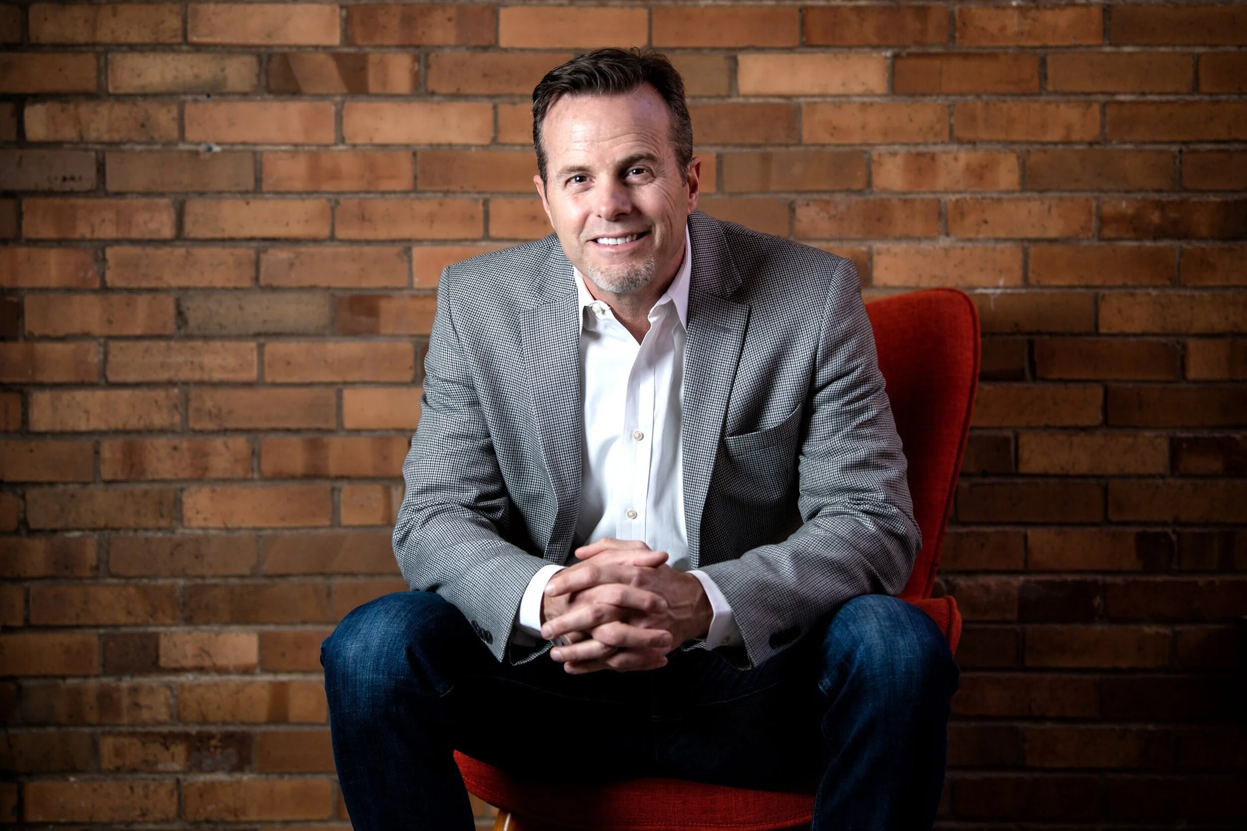 A man with brown hair and a goatee, wearing a gray checkered blazer, white shirt, and dark jeans, sitting on a red chair in front of a brick wall, smiling at the camera.