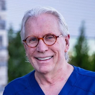 A smiling elderly man with gray hair and glasses, wearing blue medical scrubs.