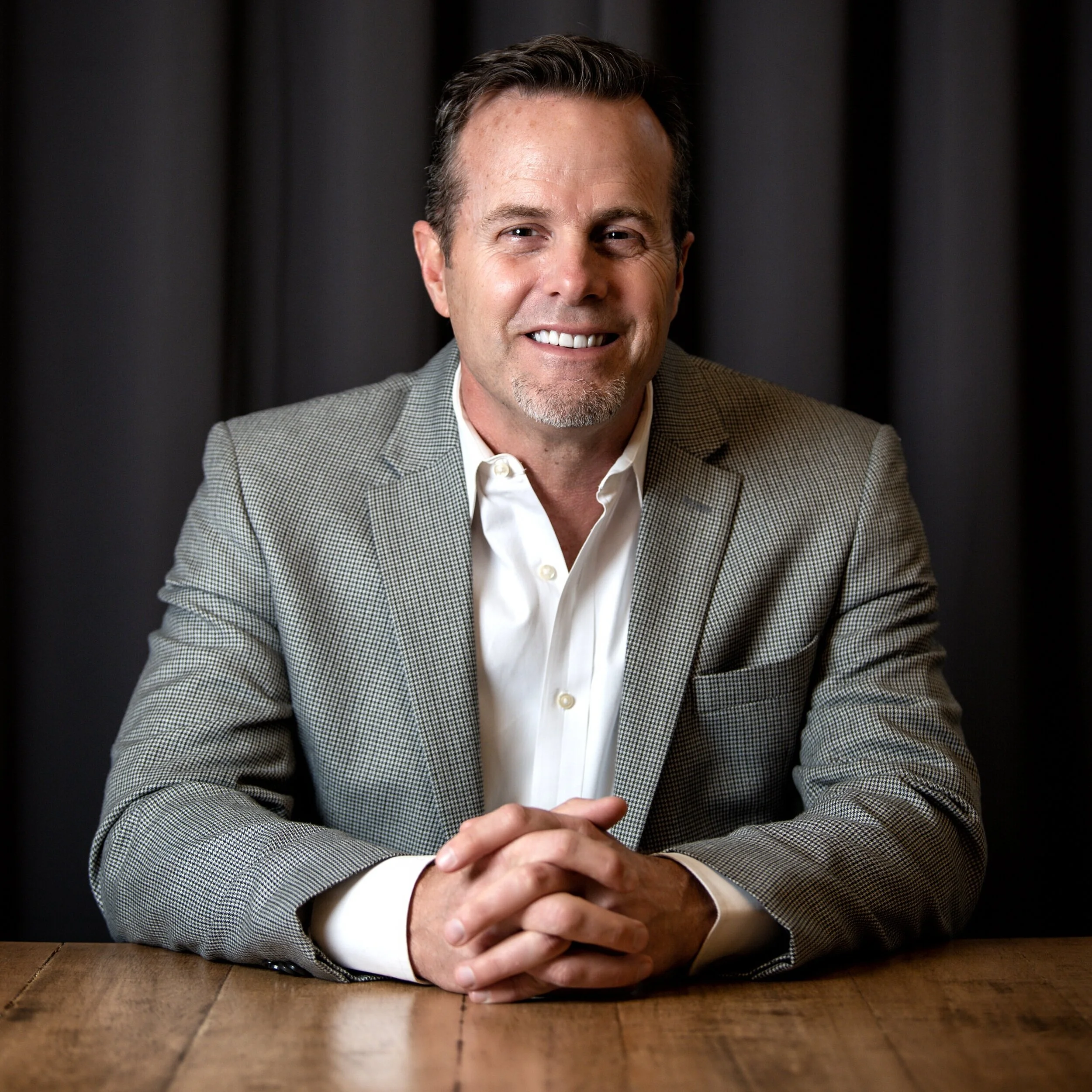A man in a light gray checkered blazer and white shirt sitting with hands clasped on a wooden table, smiling at the camera against a dark background.