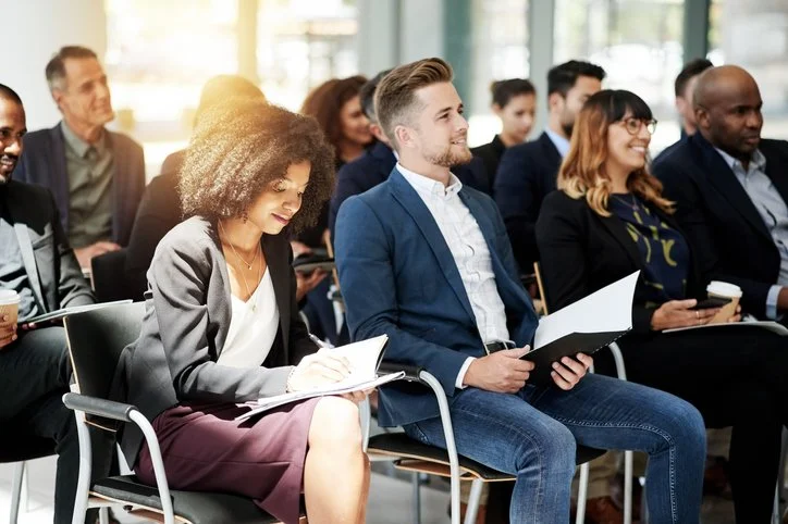 People attending a conference or seminar, sitting in chairs, with some taking notes and others listening attentively, in a well-lit room with large windows.
