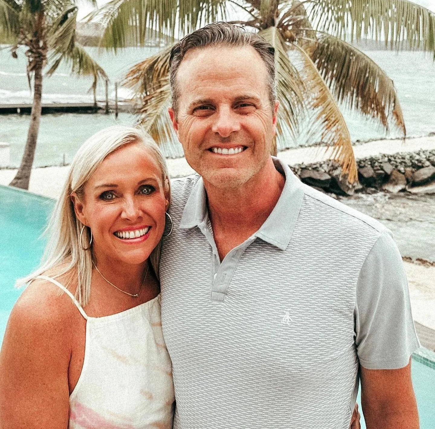 A smiling couple stands outdoors near a pool with a waterfront and palm trees in the background.