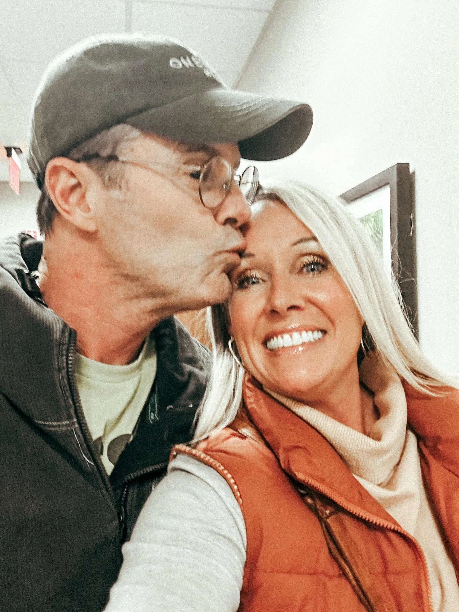 A man kissing a woman on the cheek, both smiling indoors.