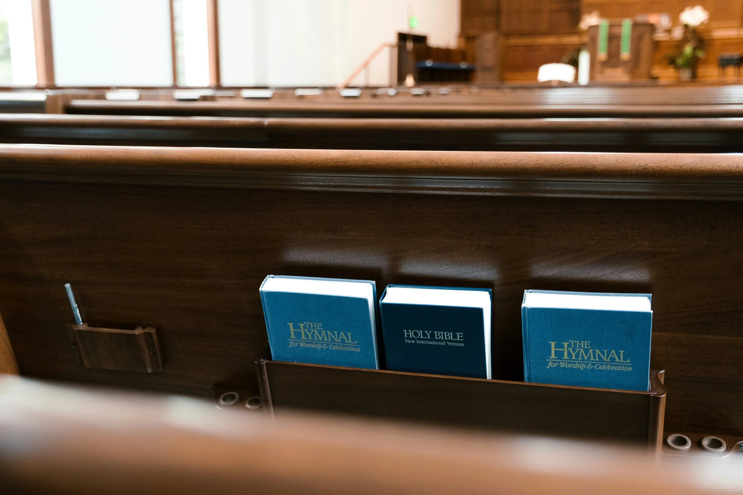 Inside a church, three hymnals including 'The Hymnal for Worship & Celebration' and the 'New International Version' Bible are placed in a wooden pew pocket, with empty pews visible in the background.