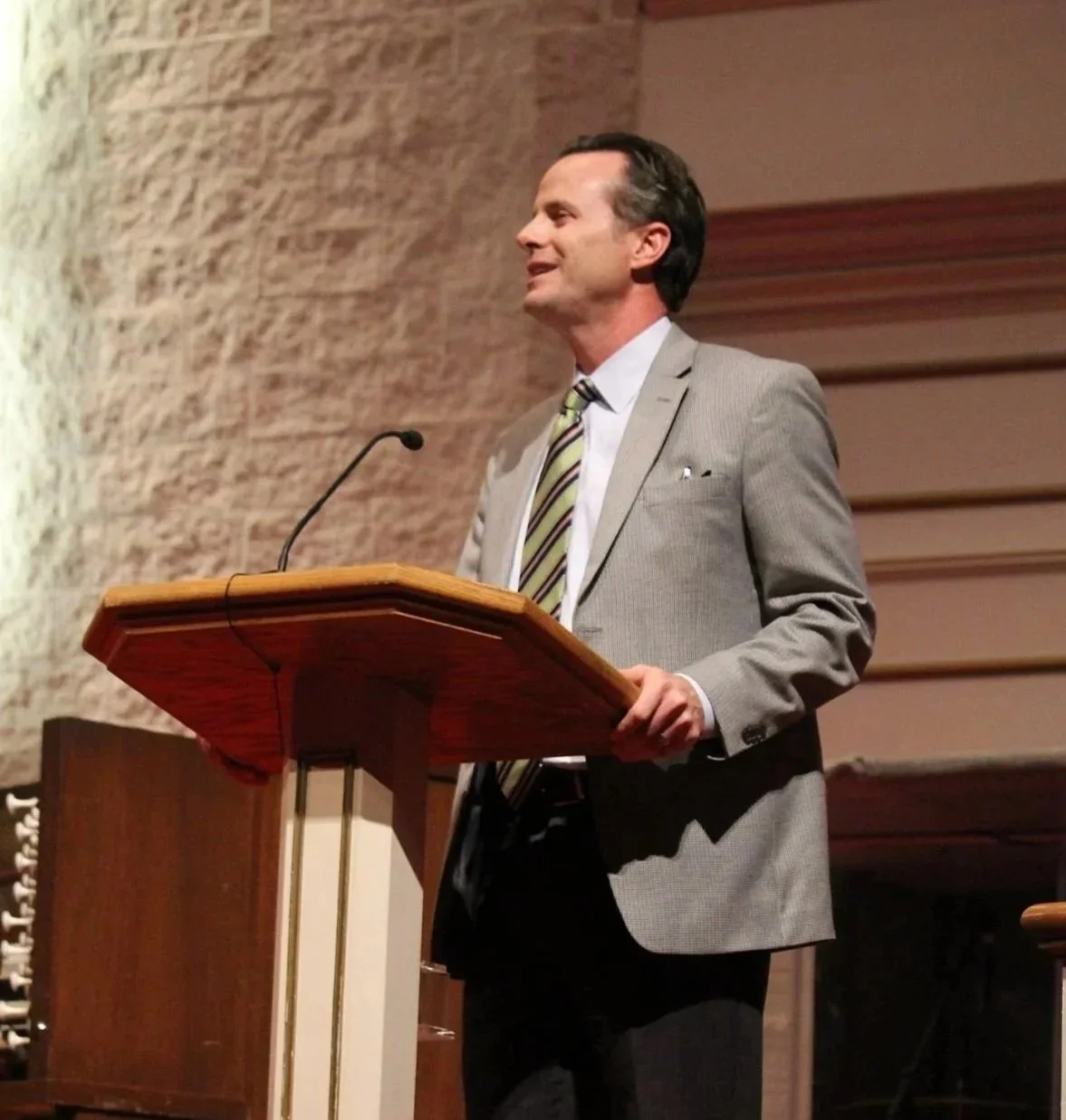 A man in a light gray suit and striped tie standing at a wooden podium, speaking into a microphone in a formal setting with brick and wood panel walls.