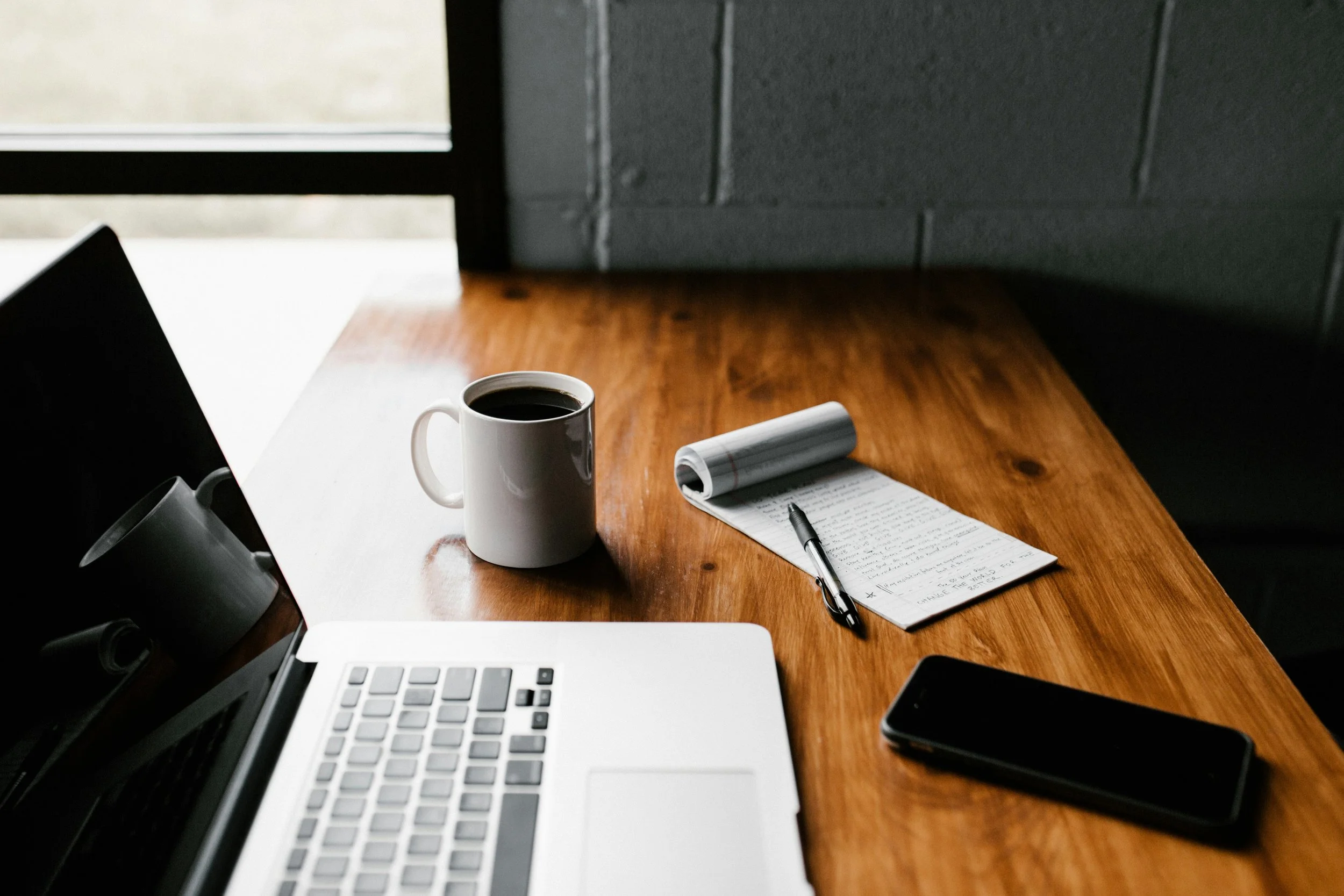 A wooden desk with a laptop, a cup of coffee, a smartphone, a pen, and a notepad with writing, in a room with a large window and gray brick wall.
