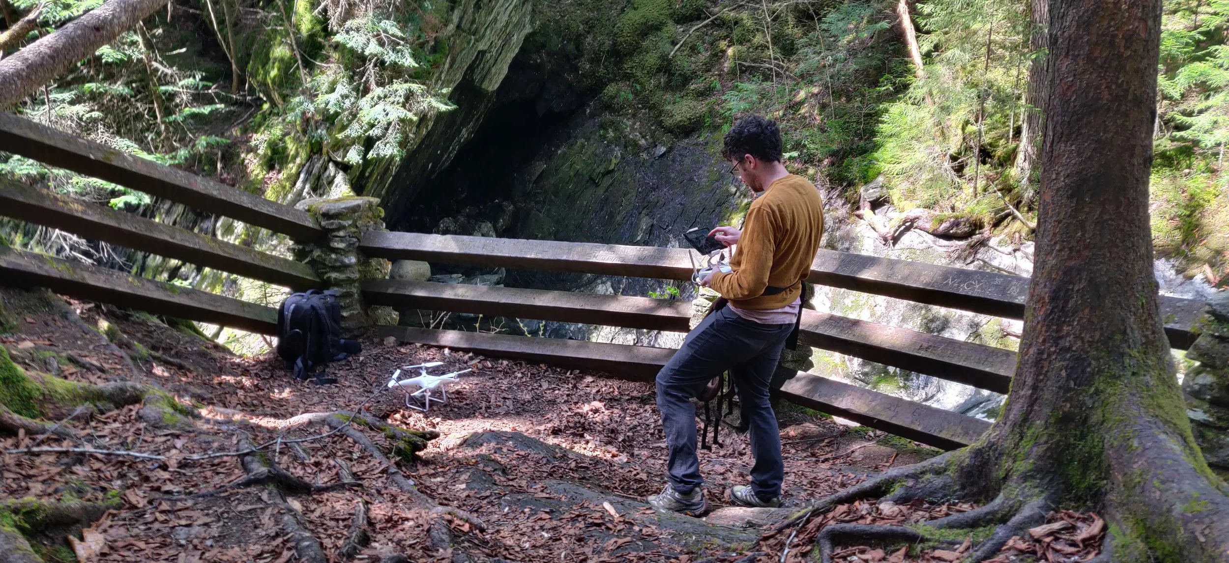 A person in a yellow sweatshirt on a wooded trail near a wooden fence, looking at their drone controller. There is a white drone on the ground and a backpack nearby, surrounded by trees and moss-covered rocks.