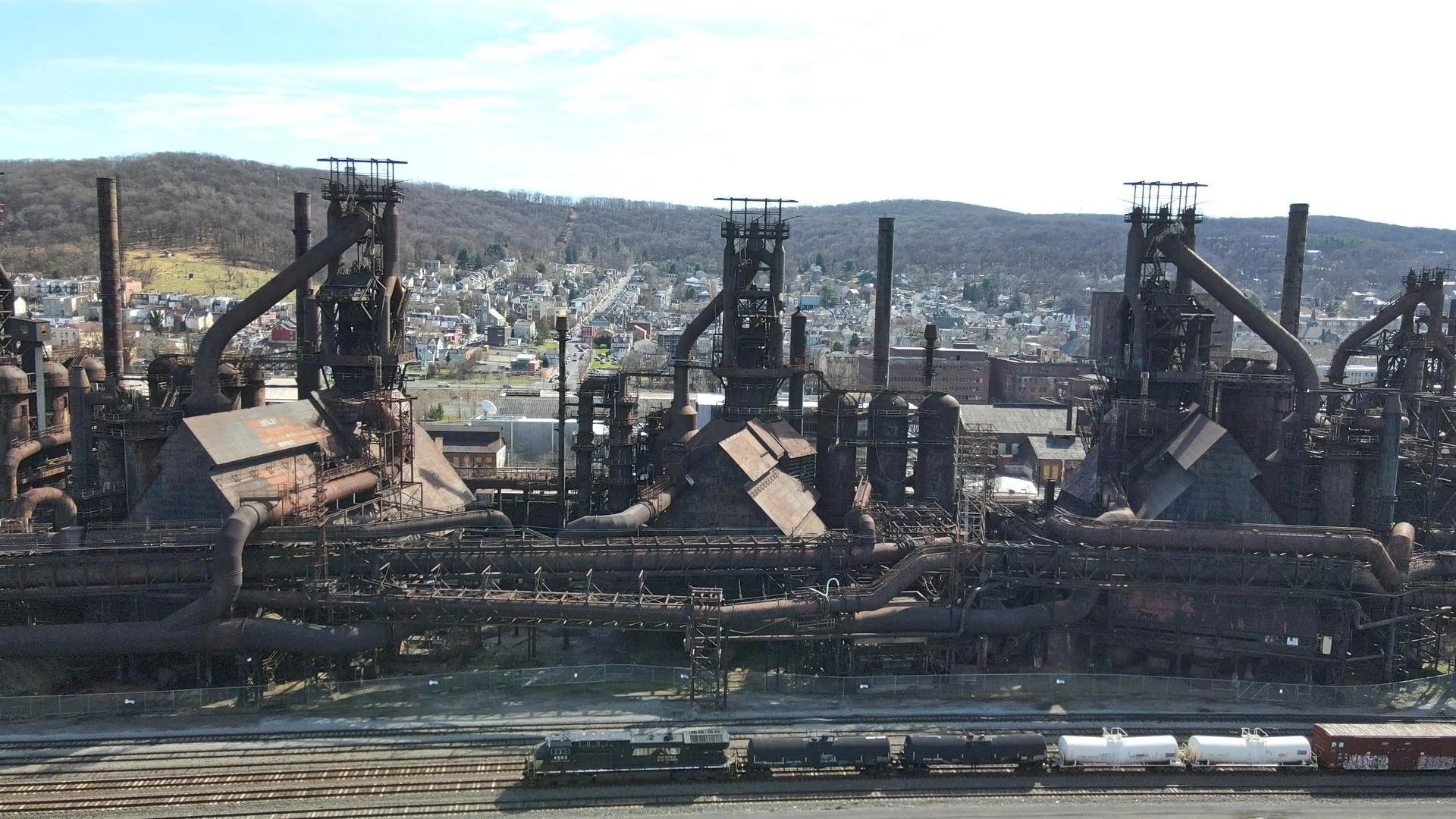 An old, rusty industrial factory with large chimneys and pipes, with a cityscape and hills in the background under a partly cloudy sky.