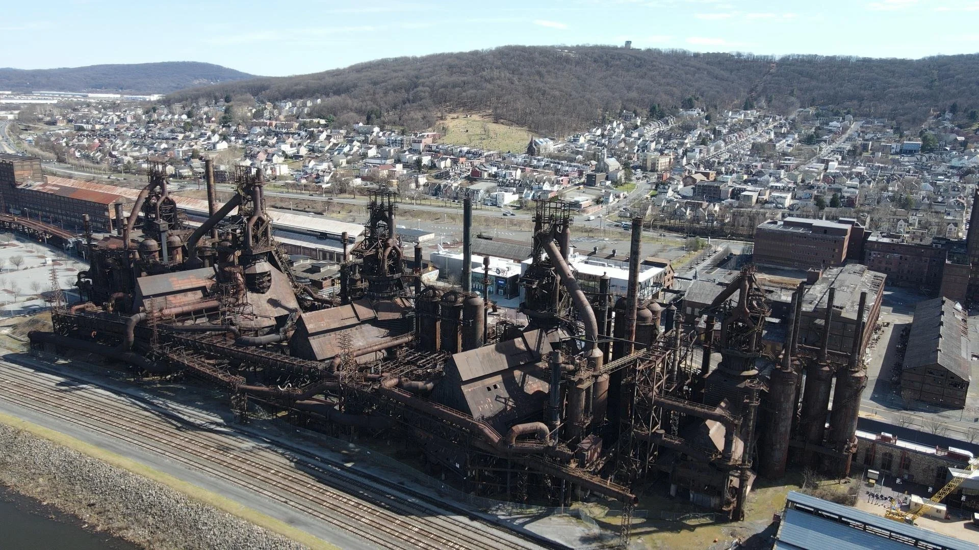 Aerial view of an old rusted industrial factory with multiple smokestacks, pipes, and buildings, surrounded by a cityscape and hilly terrain in the background.