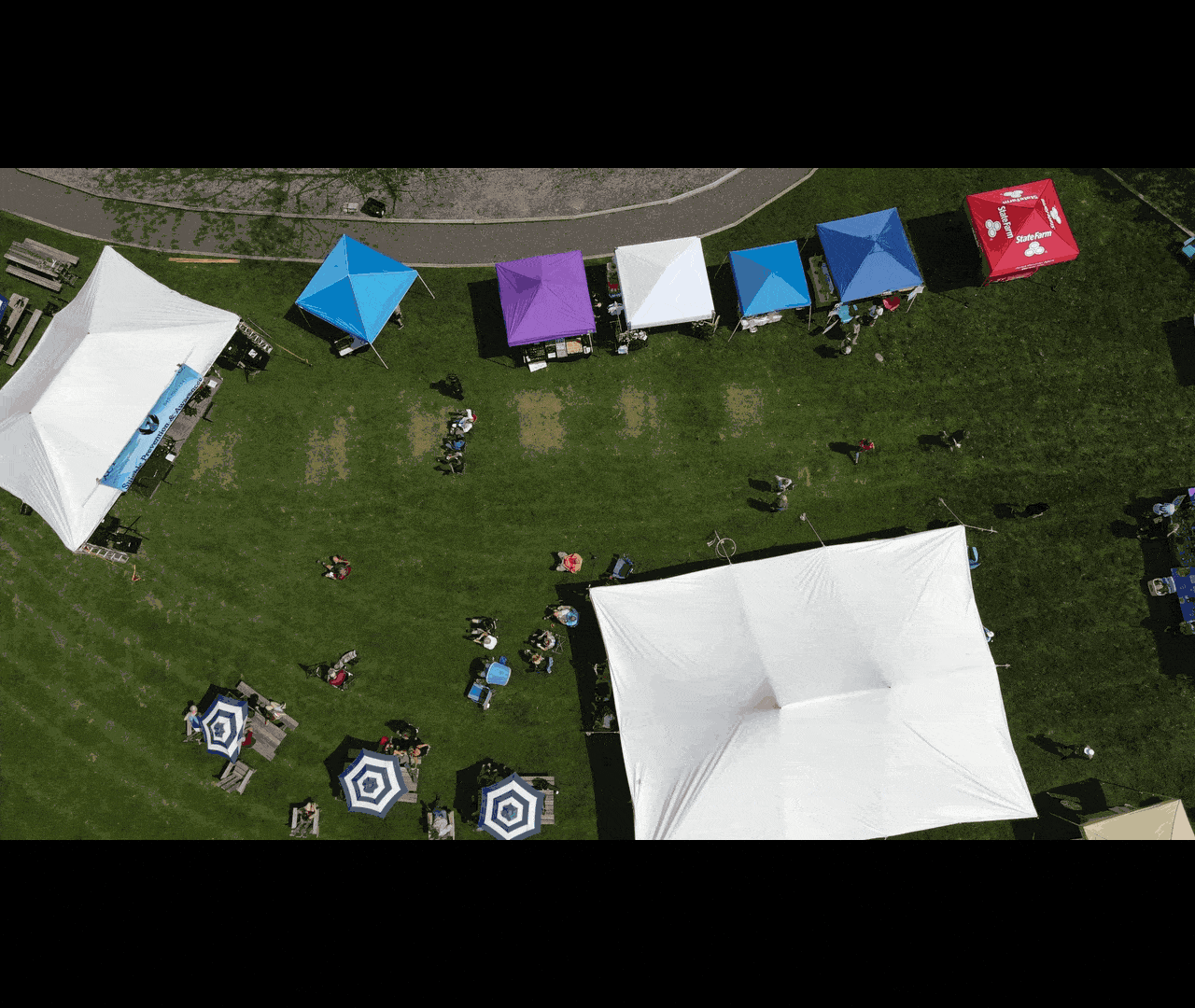 An aerial view of an outdoor event with tents, umbrellas, and people on a grassy area near a paved pathway.